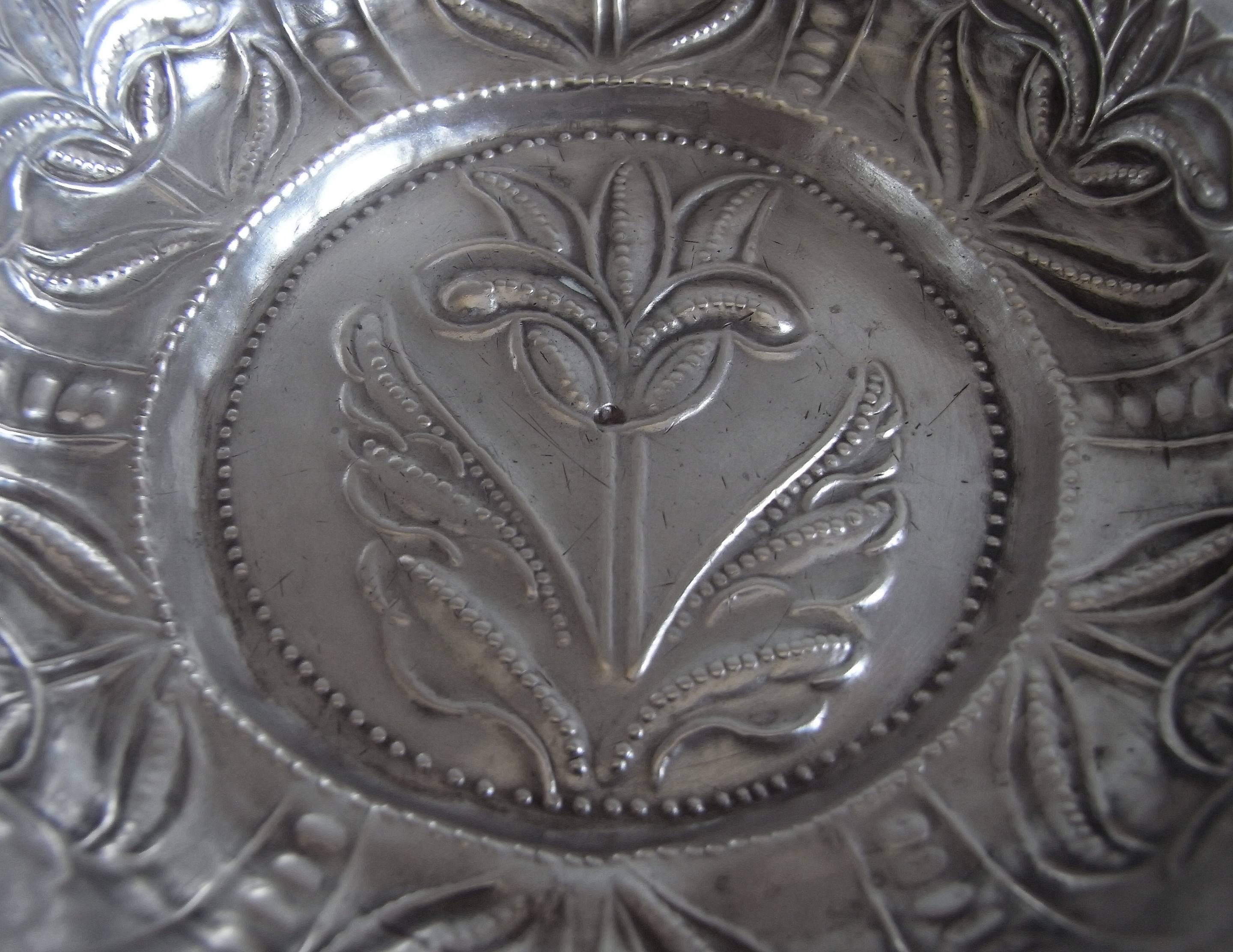 The Wine Taster is circular in form, with raised sides divided into panels by punch beaded bands, which are decorated with stylised flowers. The centre of the bowl is decorated with an unusual stylised flower head, possibly a tulip, and the base is