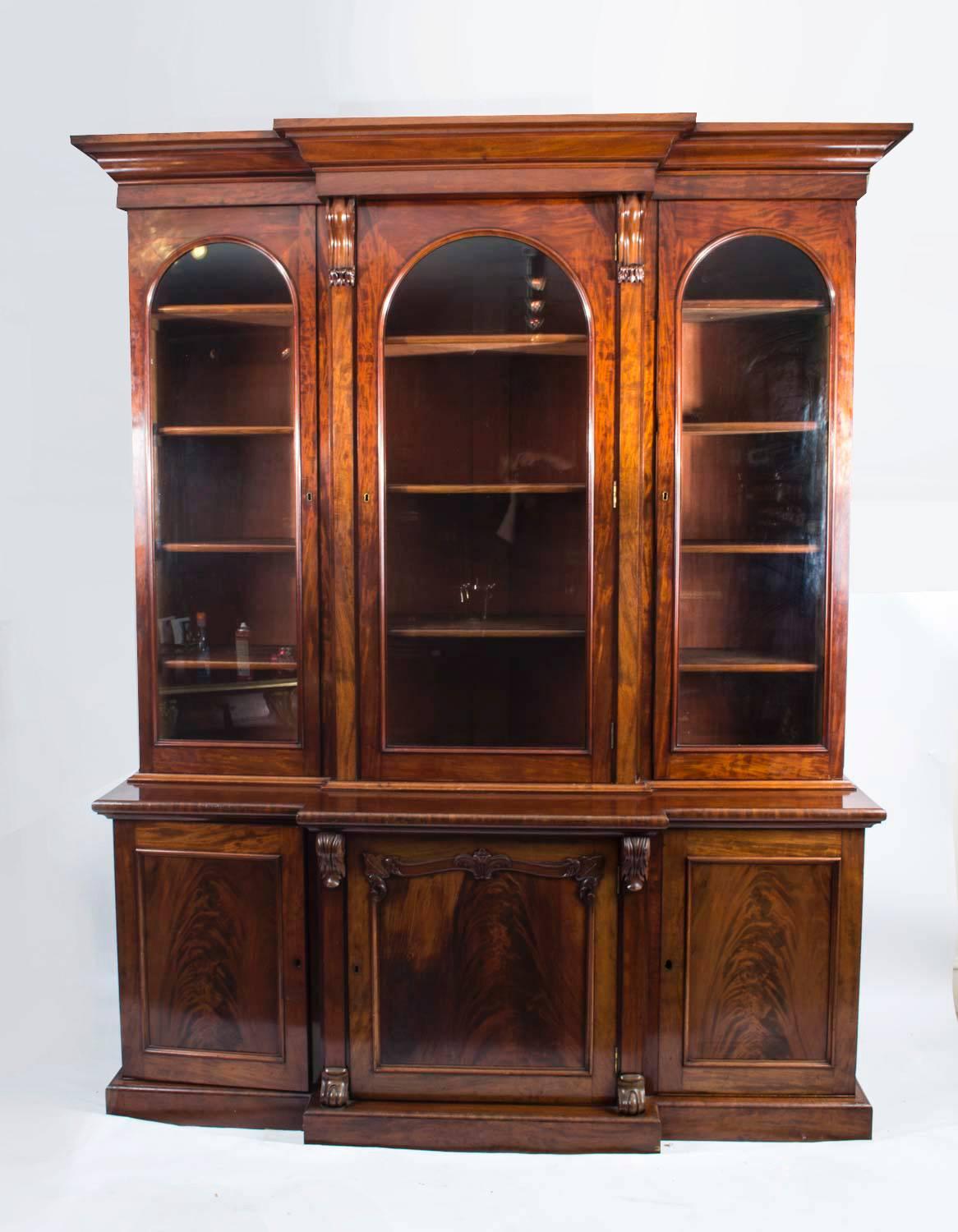 This is a beautiful antique Victorian flame mahogany breakfront bookcase, masterfully crafted in rich mahogany, circa 1860 in date. 

This grand bookcase features an upper section with three arched glazed panel doors, spaced by corbel like