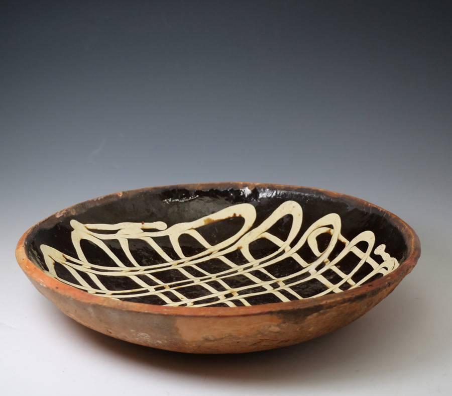 A good sized slipware baking or loaf dish circular form with strong slip decoartion on a dark chocolate ground with an elaborate and finely executed criss-cross lattice pattern. 
The deft application of the trailing slip is a testament to the