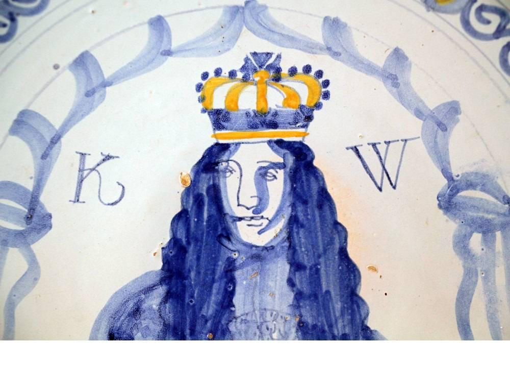 Rare English delftware pottery charger with portrait of King William 111. The bust length crowned figure is painted in blue on a soft pale blue ground and flanked by the initials WR below scrolling drapery. The distinctive border comprises