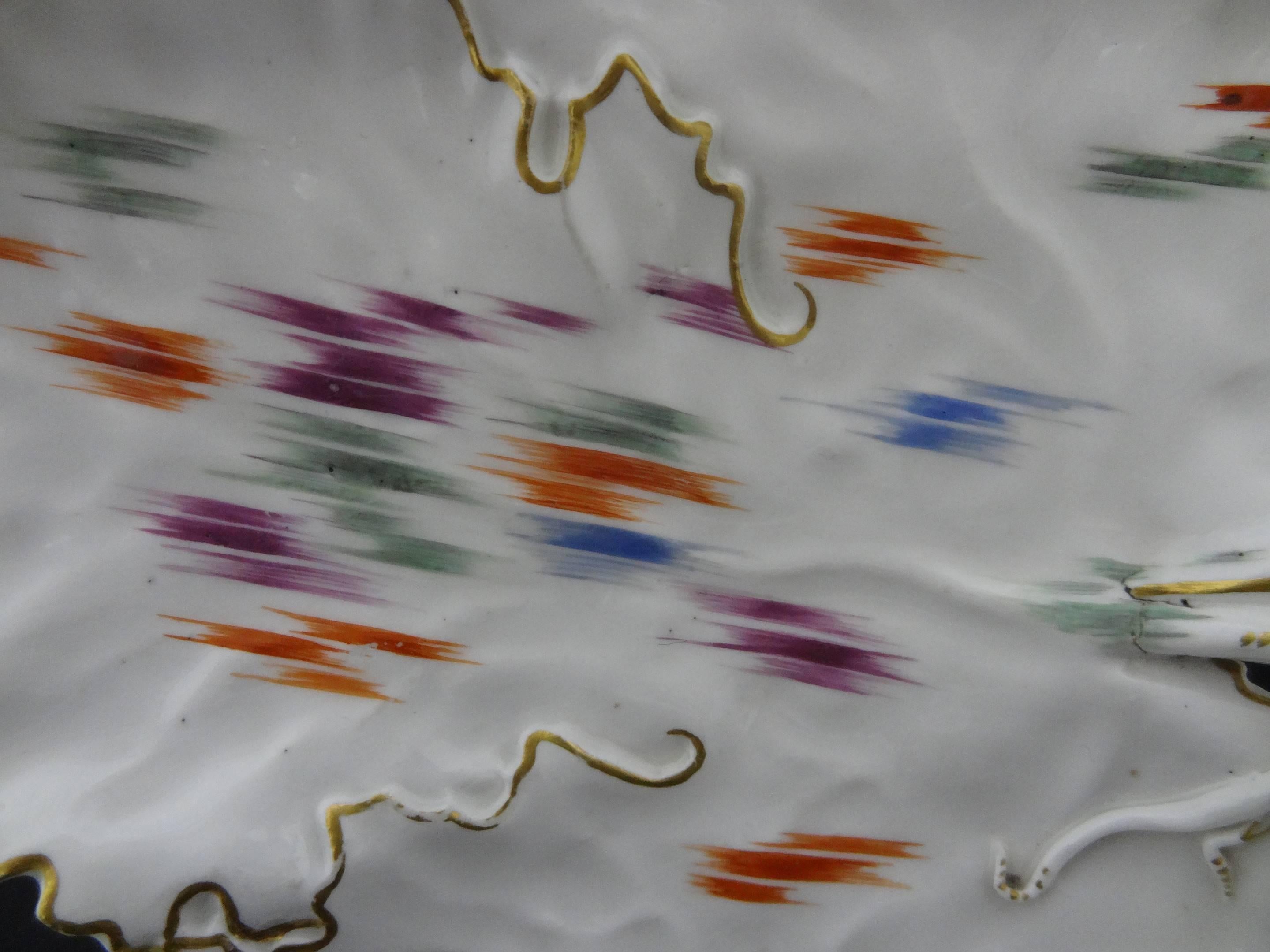 Plat à feuilles en porcelaine de Frankenthal, décoré en vert, orange, bleu et violet de courtes bandes hachurées dans des bordures de lignes dorées, couronné du monogramme CT et de 76 en bleu sous glaçure, 22 cm de long.

Ce style inhabituel de