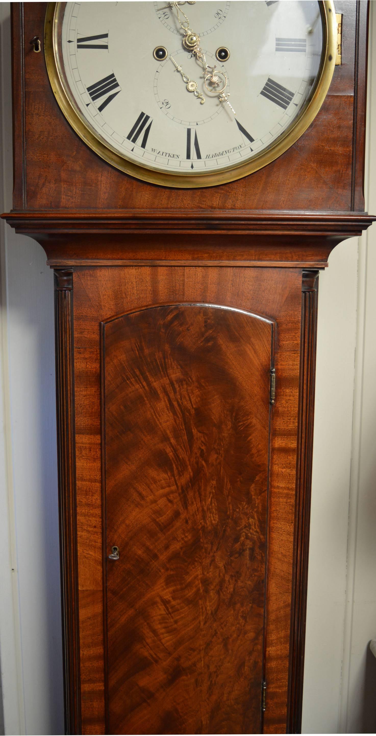 A well proportioned traditional Scottish mahogany longcase clock with round dial. The case has an arched top hood, a brass bezel and pierced sides. The trunk has a long door, with a crossbanded base , standing on bracket feet. The painted enamel
