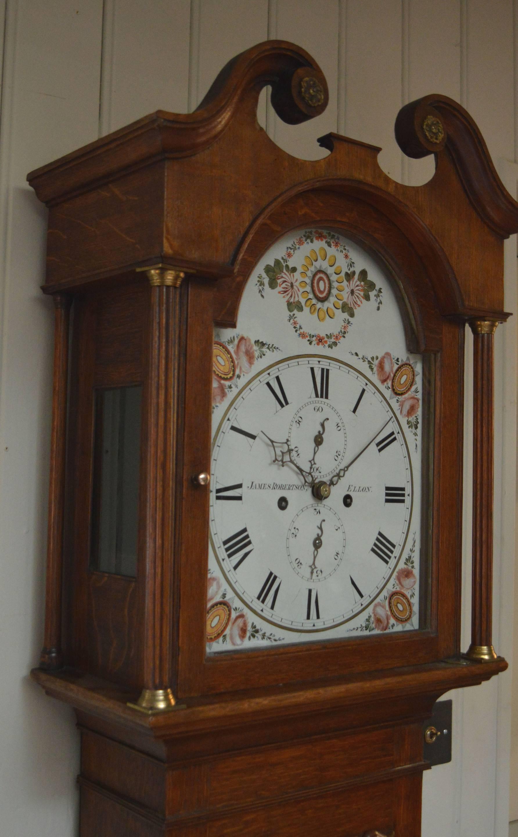 A small, light oak case 8 day longcase clock, dating to the early 19th century. It has a swan neck hood with reeded side columns and a break arched door and glass panel sides. The trunk has a long door with a shaped top. The well painted dial has