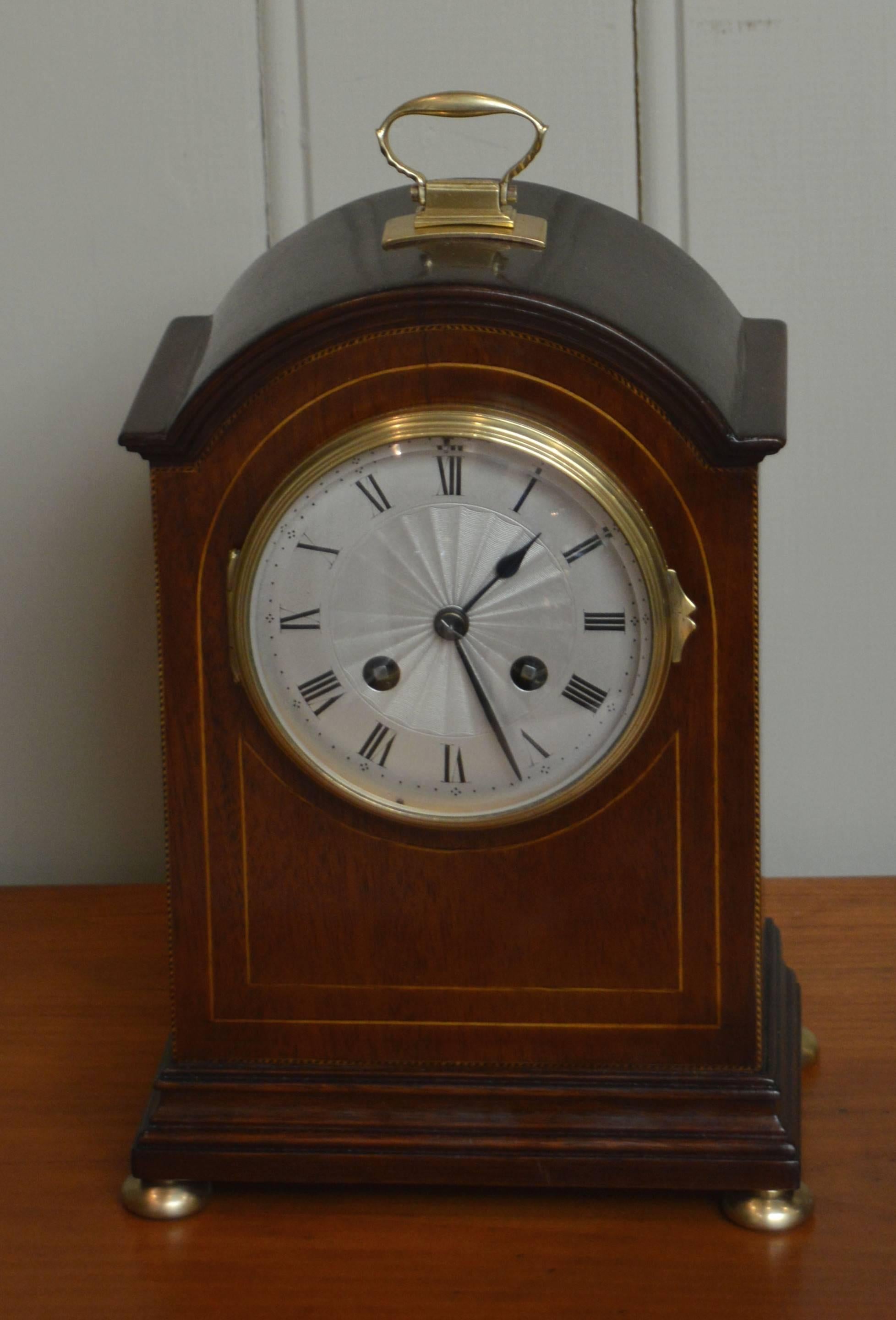 A mahogany mantel clock in the style of an English bracket clock, dating to the Edwardian period, circa 1910. It has a dome top with a brass carrying handle. The front panel has boxwood stringing and a convex glass with a silvered engine turned