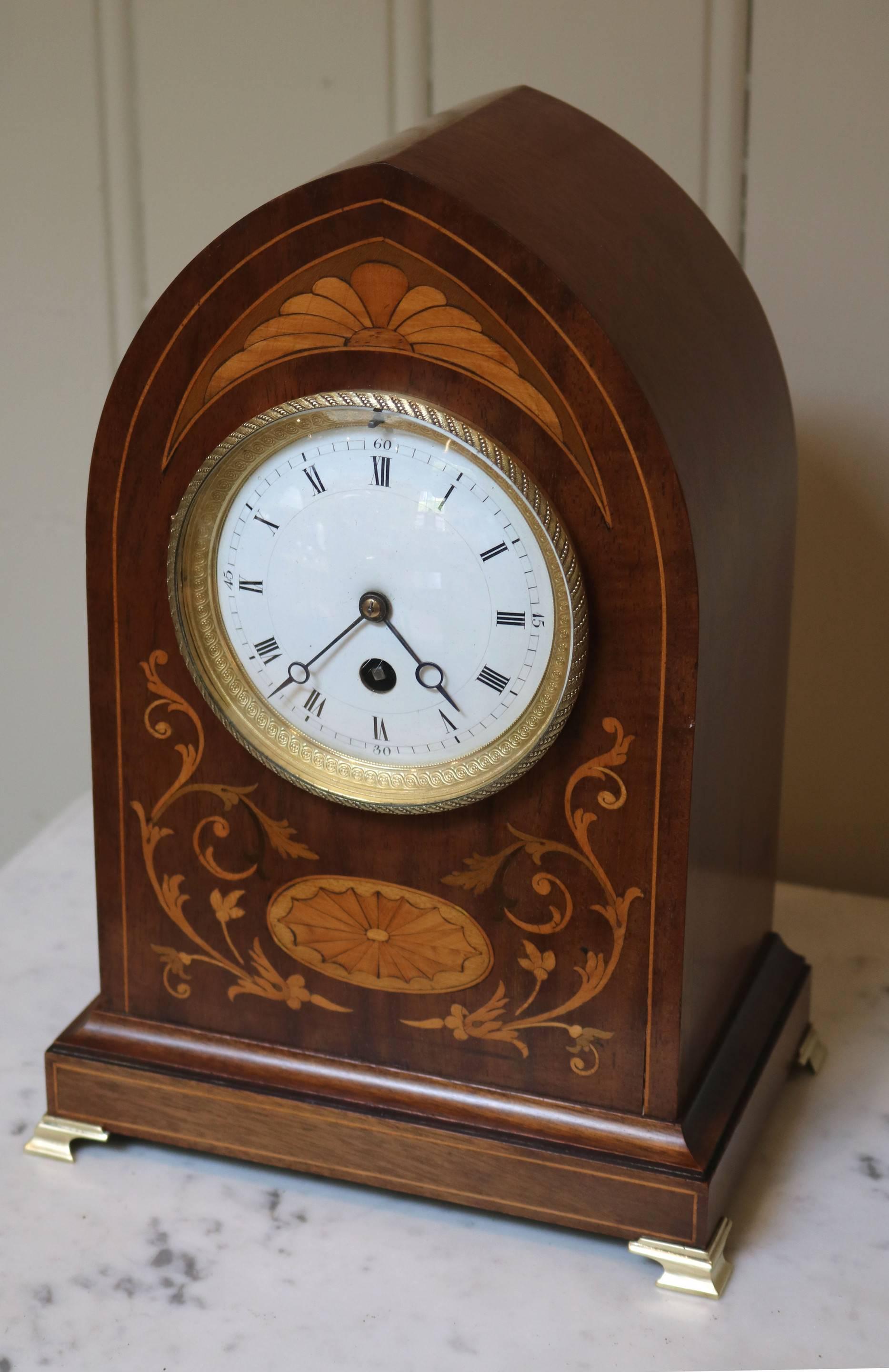 A mahogany lancet top mantel clock. It has a lancet top case with a front panel inlaid with boxwood stringing and floral marquetry satinwood inlays. It has a convex glass with an embossed bezel and sight ring and white enamel dial with steel moon