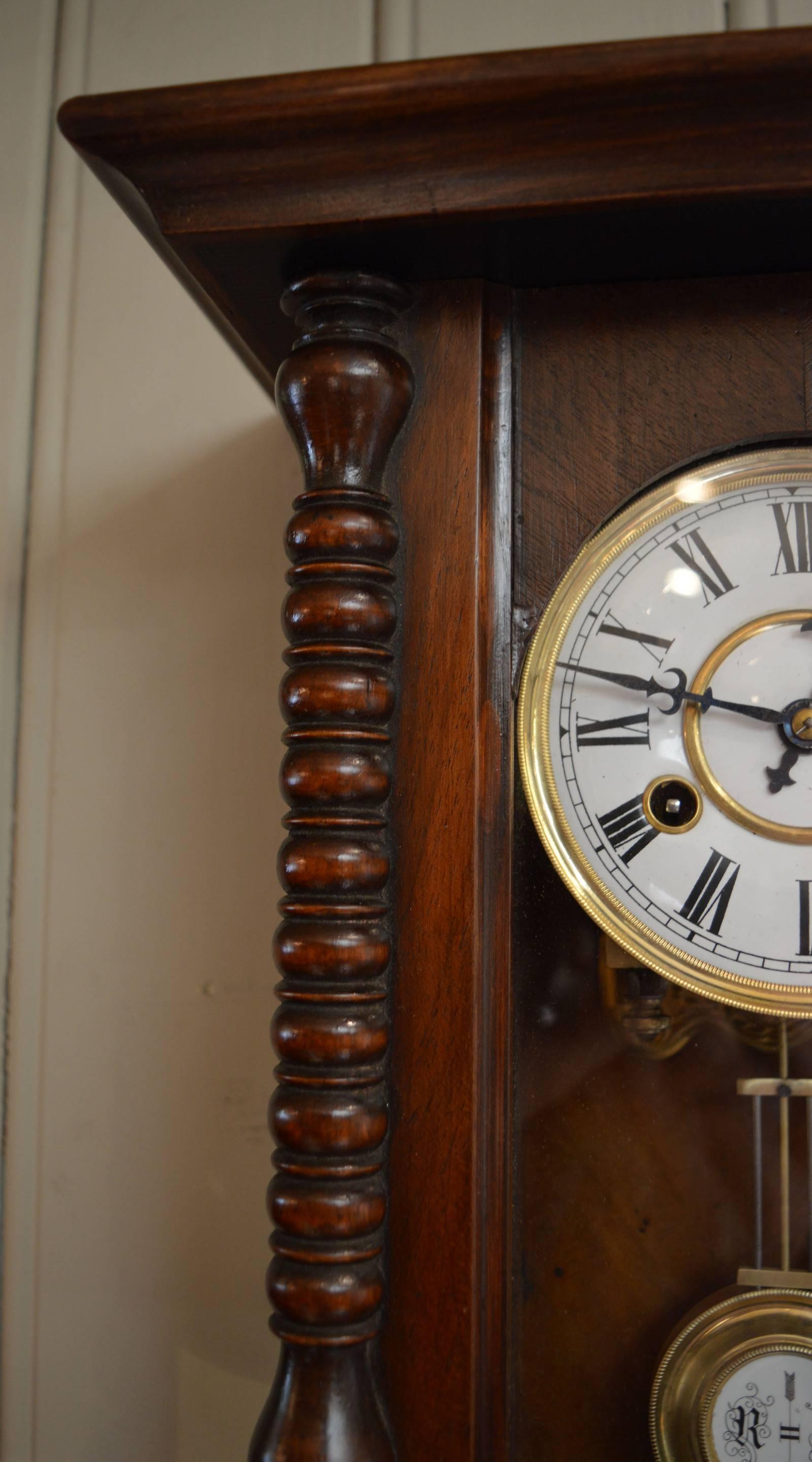 A small, late 19th century striking walnut wall clock. It has a regulator style case with turned bobbin columns and a shaped base. The two-piece enamel dial has Roman numerals and a matching simulated gridiron pendulum with retard and advance and