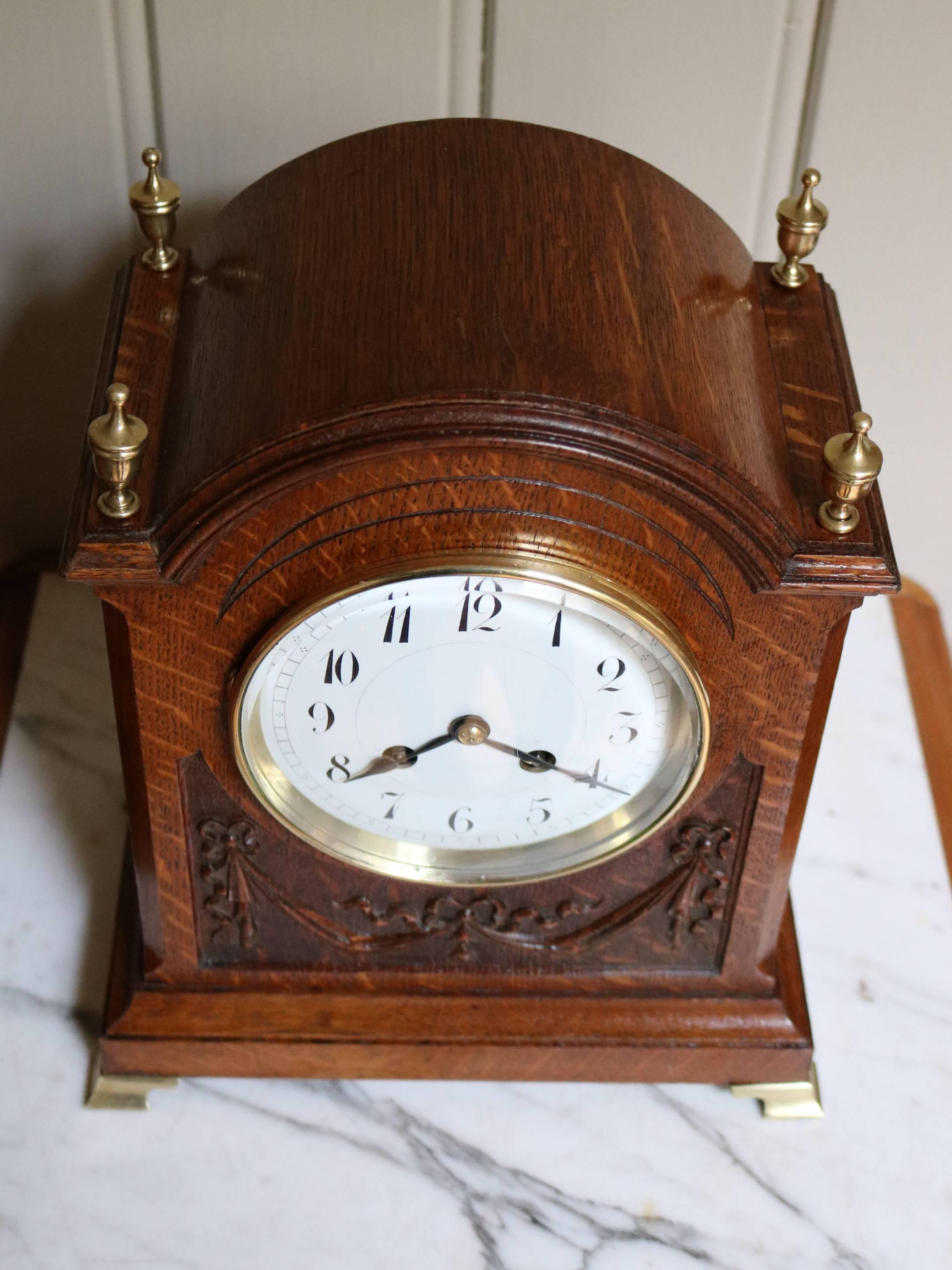An attractive solid oak mantel clock dating to the early 20th century. It has an arch top with four brass corner finials, a well carved central panel of swags and ribbons,a stepped base standing on brass bracket feet. The enamel dial has a brass