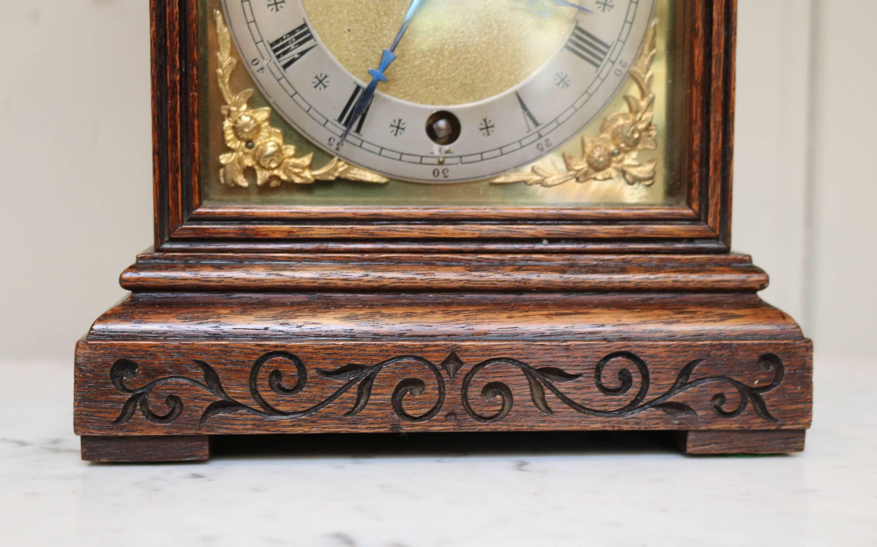 A small, solid oak case mantel clock dating to the early 20th century. It has an arch top with scroll engraving and a square dial. The brass dial has a silvered chapter ring with a matted centre and cherub spandrels. The eight day timepiece pendulum