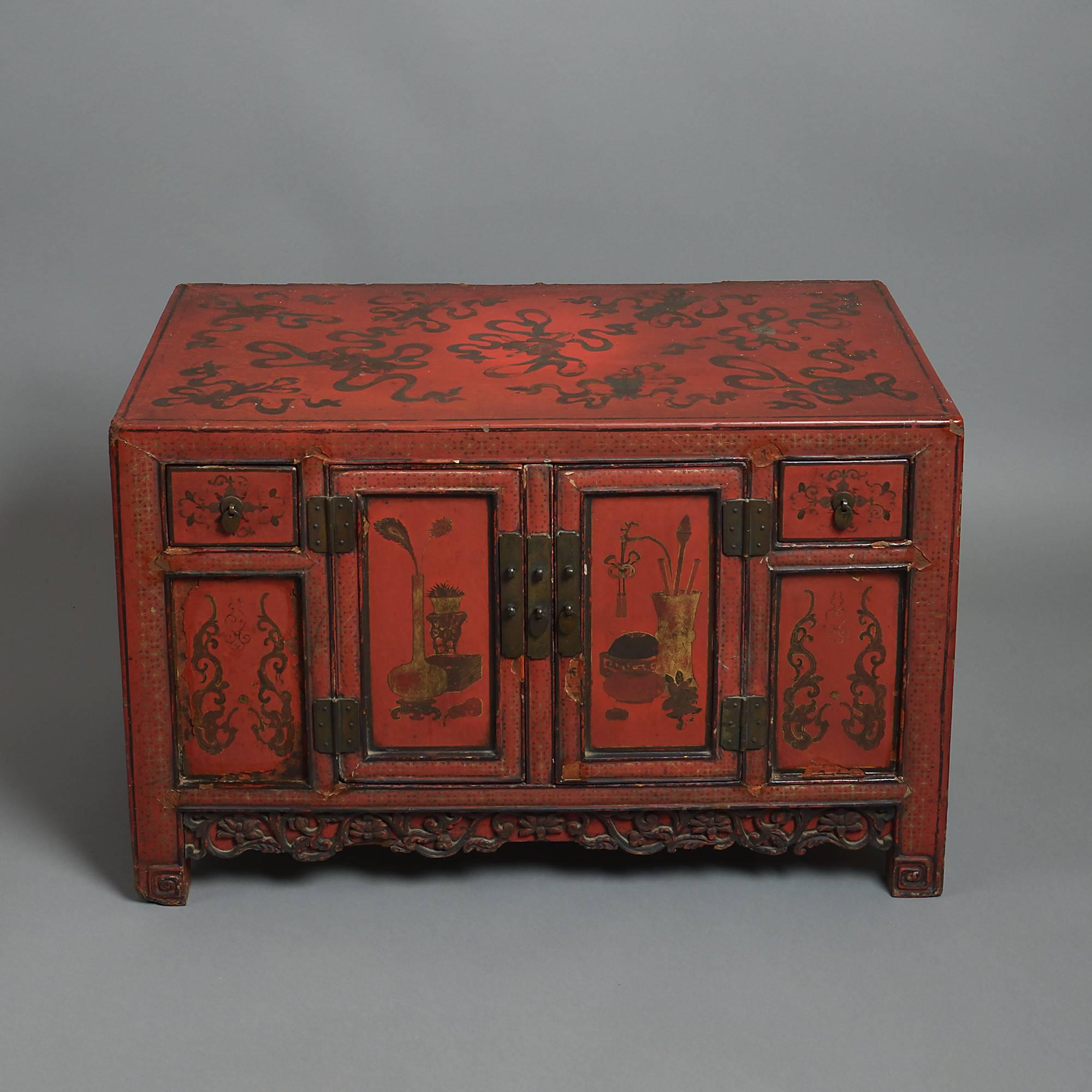 A pair of 19th century red lacquer low cabinets having each double doors and two short drawers above a carved apron and scrolling stile feet, the surfaces profusely decorated with stylised floral and foliate designs, the central panels with vases