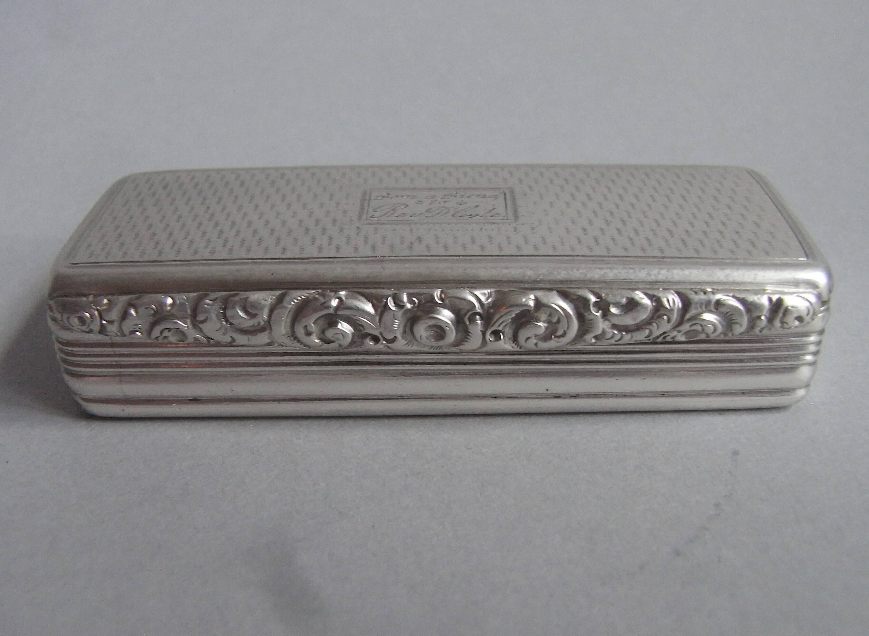 The snuff box is of slender broad rectangular form. The cover and base are decorated with engine turned designs and the sides with reeding. This example displays a floral and foliate cast thumb piece and the cover also displays an oblong cartouche