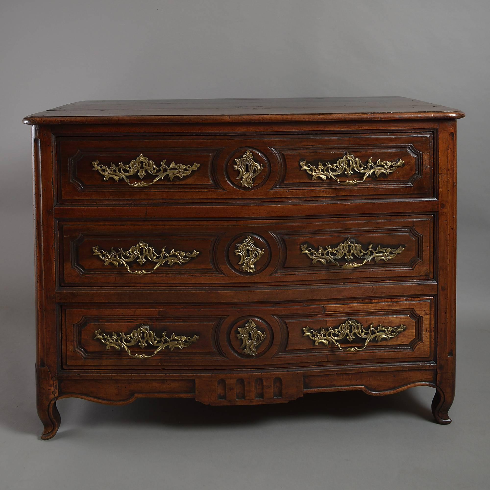 An early 18th century walnut commode, the overhanging top above three panelled drawers, each with later finely chased brass Rococo handles, the sides having arched panels, all set upon a shaped apron terminating in scrolling feet.