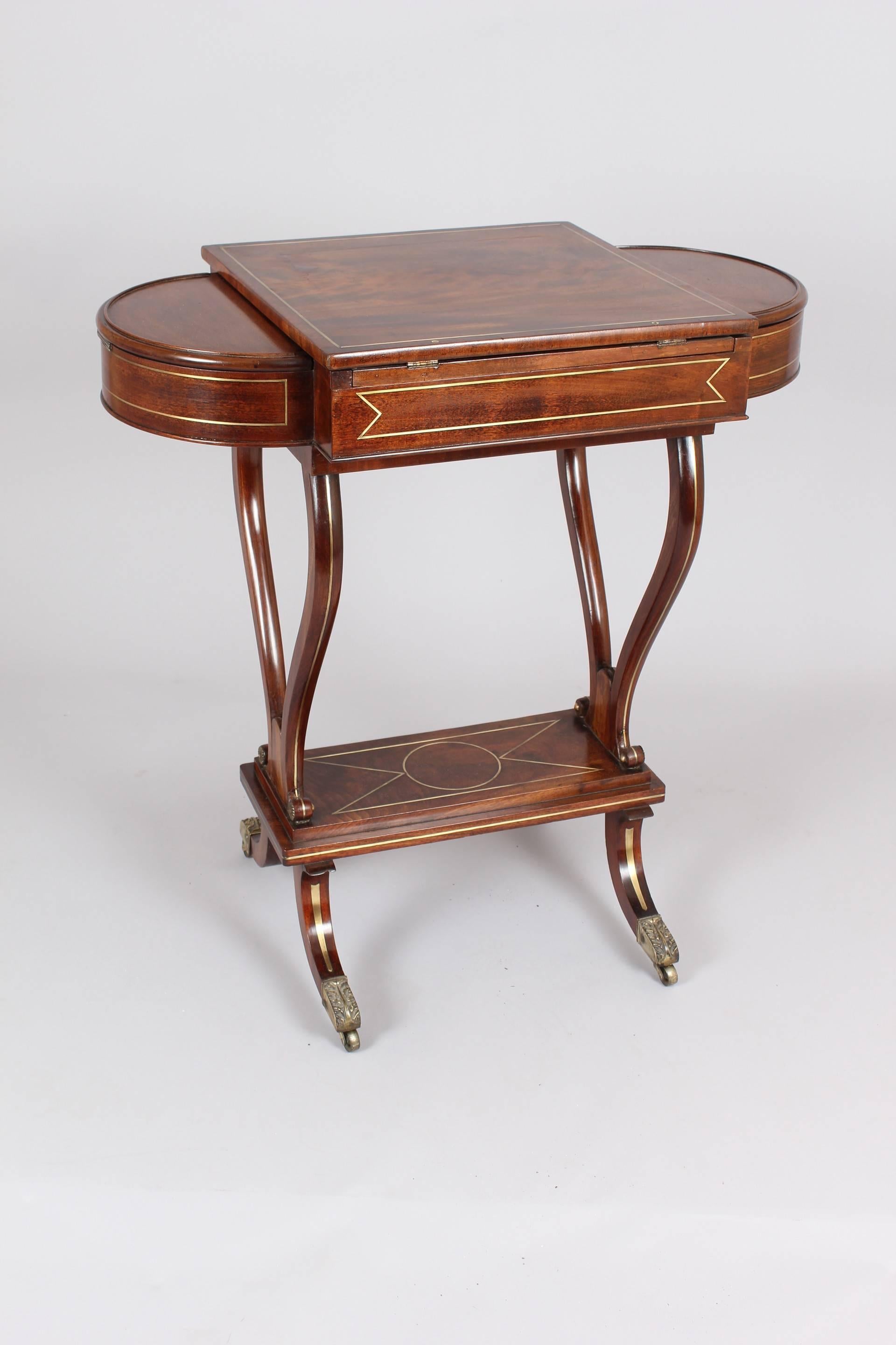 Regency mahogany and brass inlaid work-table; the hinged and adjustable top sliding forward to reveal a multi-compartmented interior and flanked by semi-circular boxes with hinged lids; on scrolled end-supports and a central platform with sabre legs