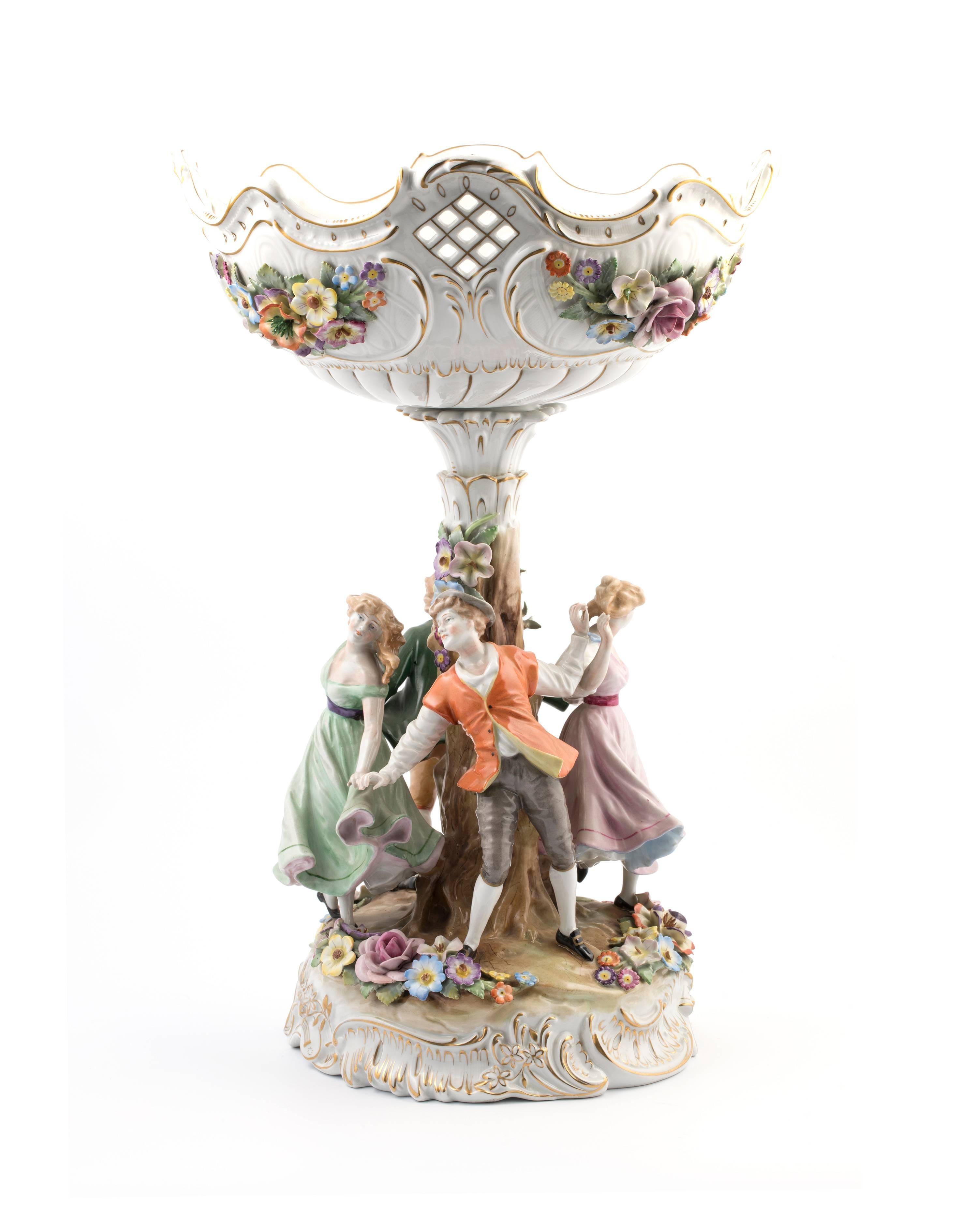 Porcelain centrepiece or compote. The compote consists of a reticulated round bowl with scalloped edge and profuse floral encrustation. The bowl fits onto a central stylized tree like column that is surrounded by four dancing boys and girls.
On a