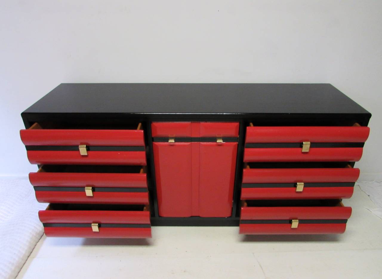 Striking American of Martinsville credenza with three drawers on either side of a swinging door that opens to reveal three dove-tailed drawers (nine drawers total).  Newly lacquered in a two-toned black and red high-gloss finish.  Original brass