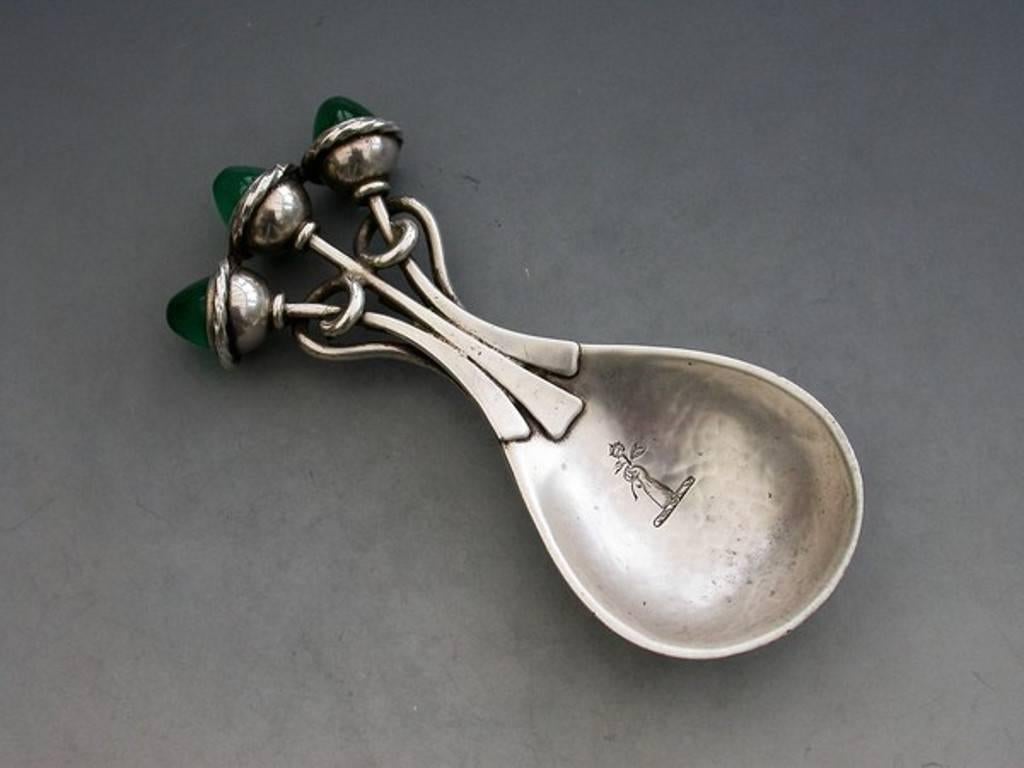An Arts & Crafts cast silver caddy spoon with hammered pear shaped bowl, the scrolling wirework stem set with three green chrysoprase cabochons on ropetwist finials. The bowl engraved with a crest - a dexter hand ppr., holding a rose gu.,