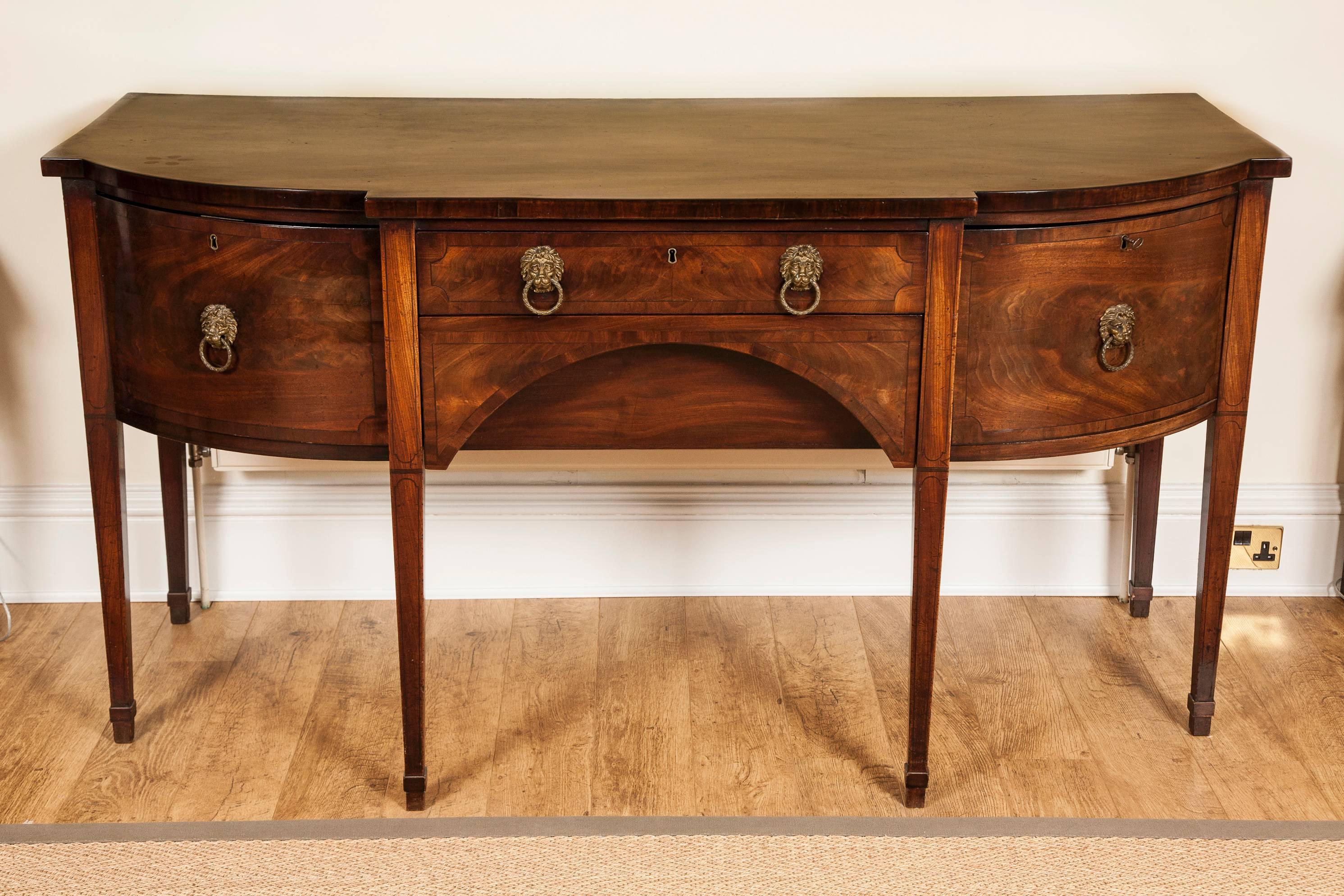 The overhanging top with bow re-entrant corners above two central and two cellarette drawers with lion mask handles, ebony stringing and mahogany crossbanding; standing on six square section tapering legs also with ebony stringing detail.
 