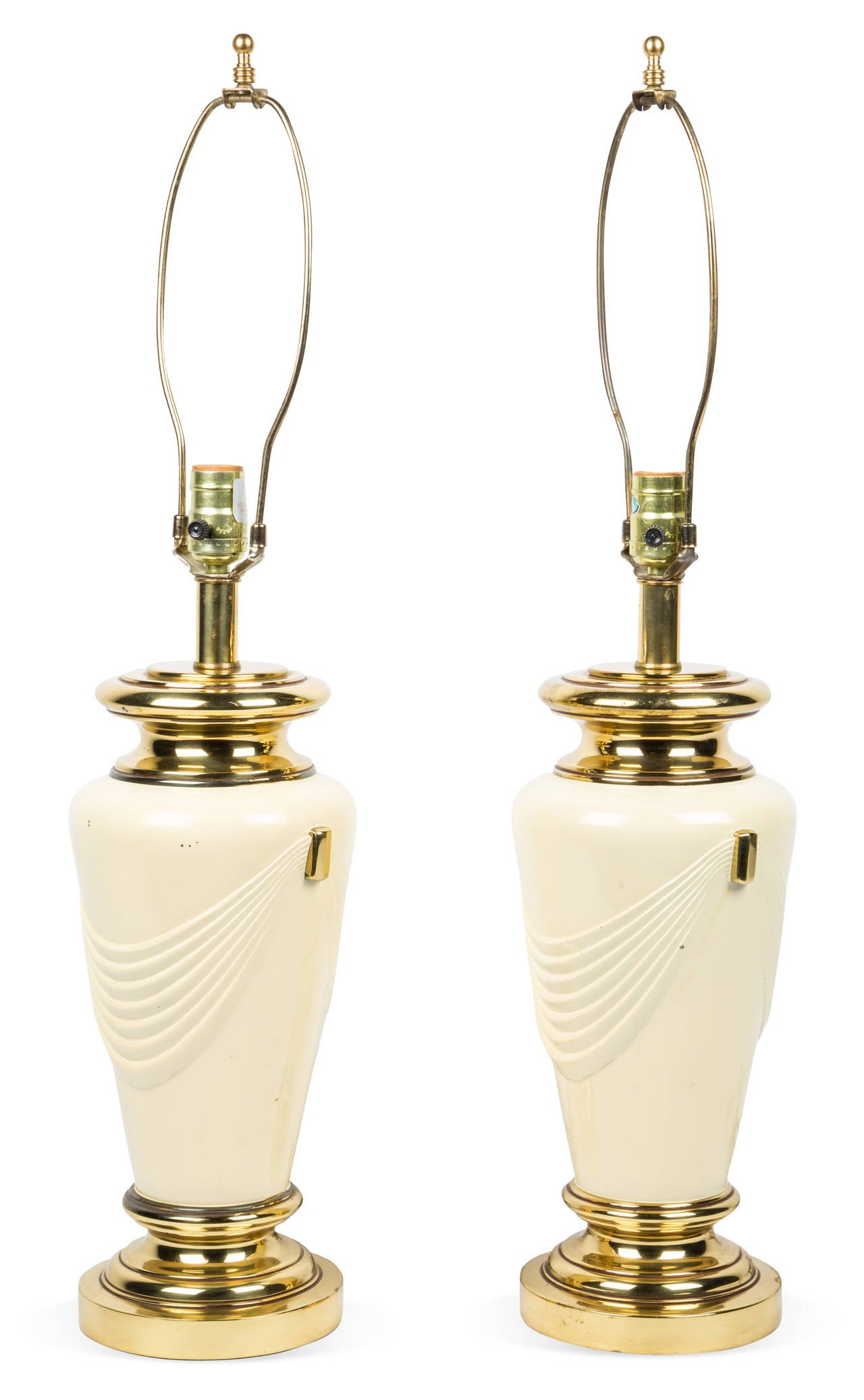 Pair of Neoclassical style brass table lamps with dimensional drape design. Base features brass details with cream gloss painted finish. Standard bulb. Original wiring in working condition. Harps and finials included.