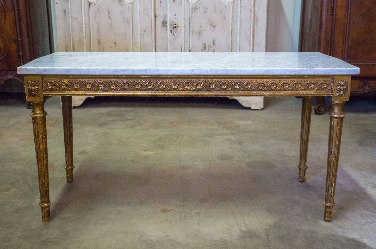 French gilded coffee table with Carrara marble top in original unrestored condition. Classic Louis XVI style with rosettes over tapered fluted legs and lovely carved apron. Mixes with other periods seamlessly.
