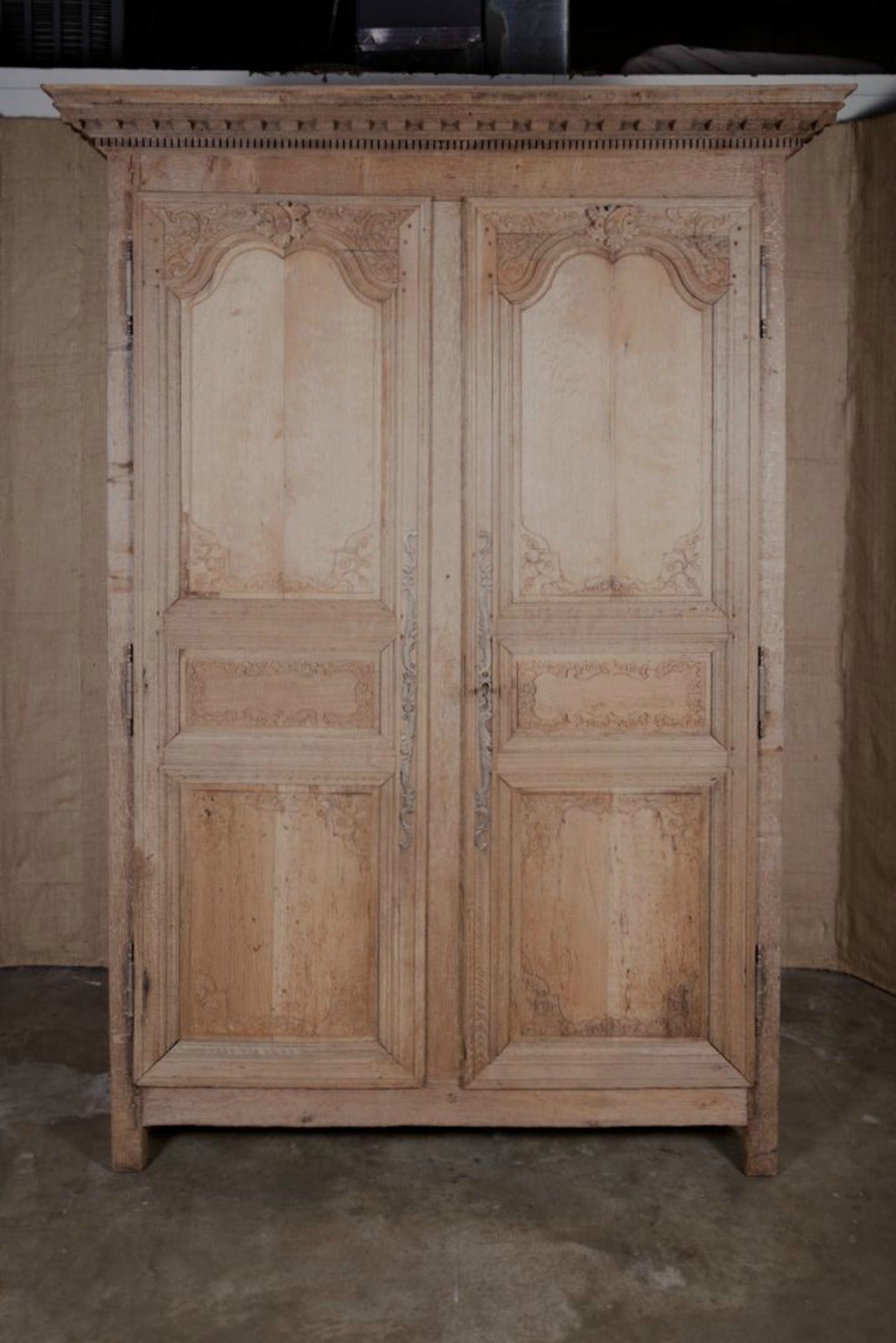 Late 18th century Country French washed oak armoire handcrafted by rural artisans, featuring timeless Louis XIV inspired motifs. The intricately carved crown with rows of dentil molding, sits above a pair of paneled doors with floral and foliate