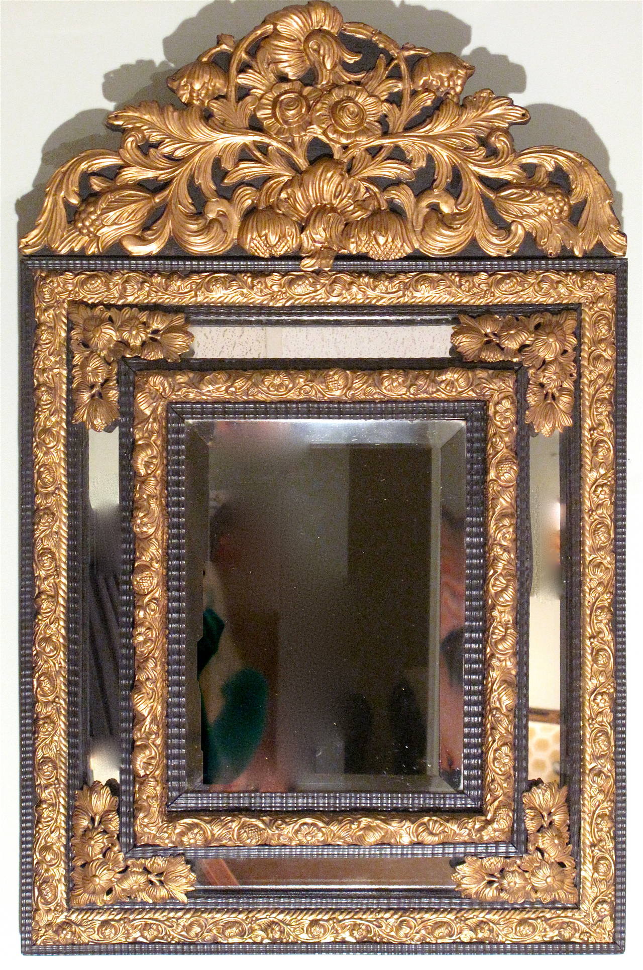A stylish Continental ebonized and gilt brass repoussé crested cushion mirror, 19th century. The arched crest is composed of bold foliate and floral elements, the layered ripple carved frame accented with band of gadrooned and foliate repoussé.