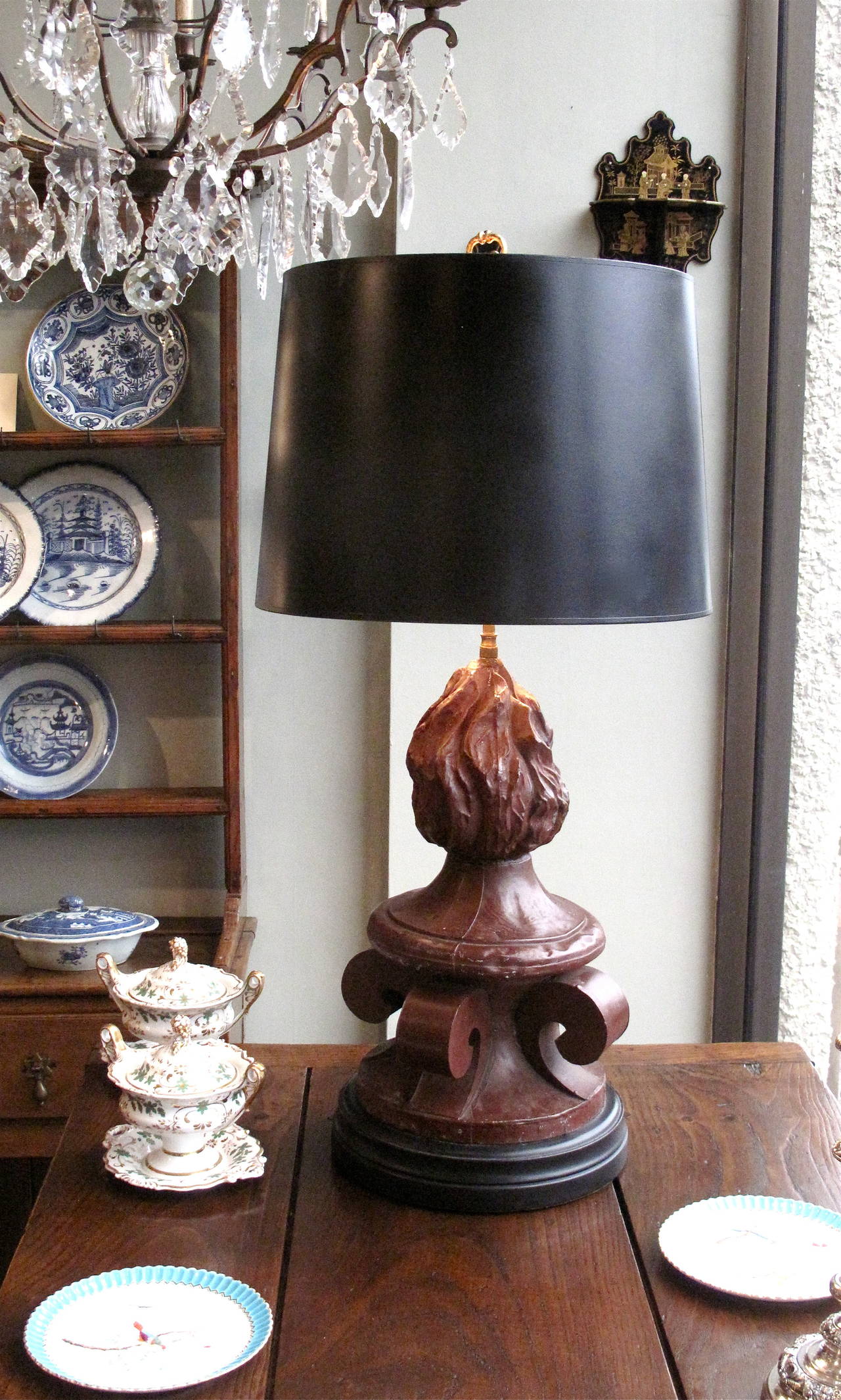 A Continental painted tôle repoussé architectural finial consisting of four volutes surmounted by a flame, later wired as a lamp, the whole resting on a wooden base. Black painted parchment drum shade.