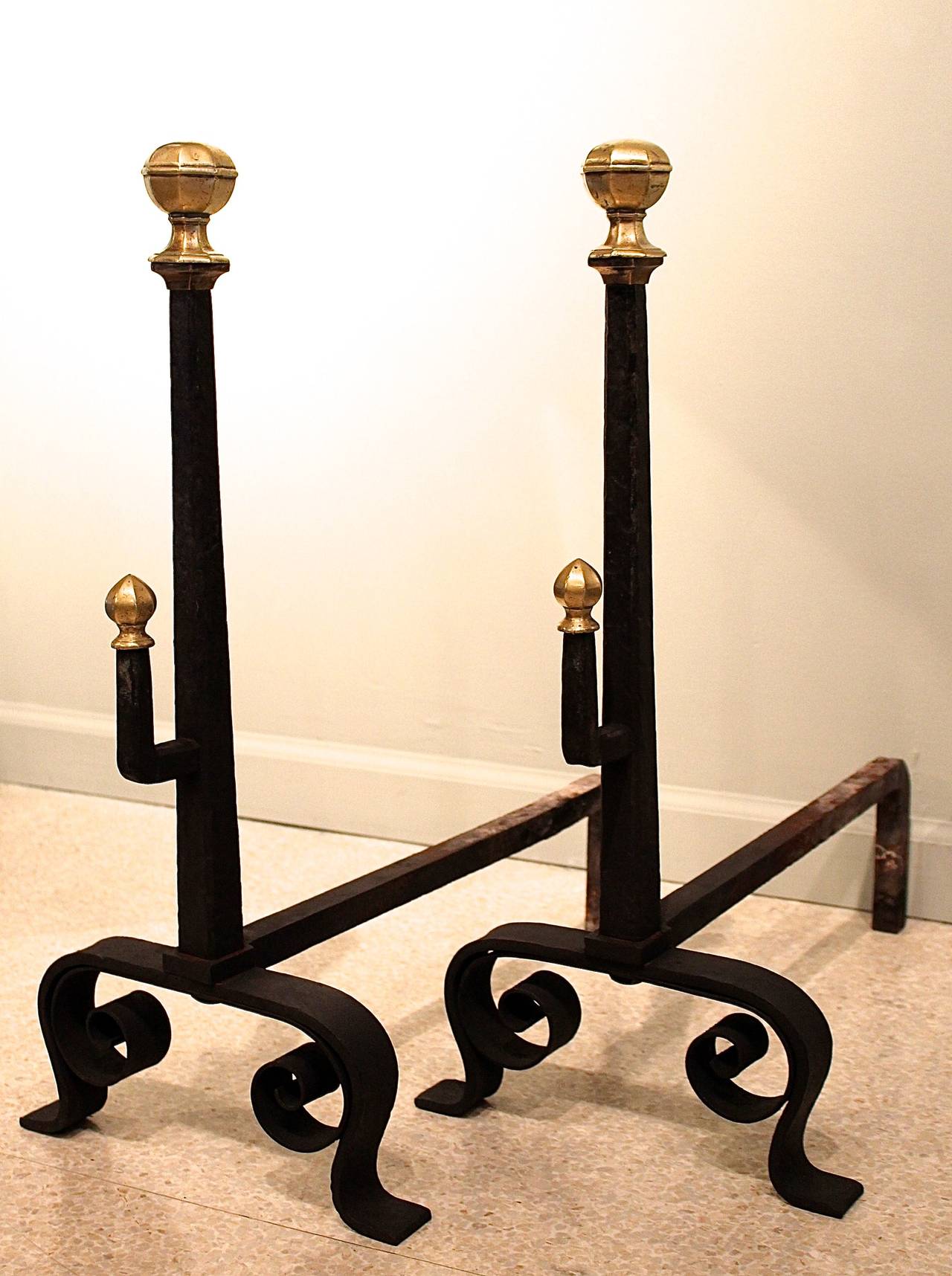 A fine pair of ca. 1900 English Arts and Crafts era andirons of wrought iron, the guards resting on scroll feet and with hooks in front for holding a roasting spit or other implements. Attractive faceted and moulded finials top the guards and hooks.