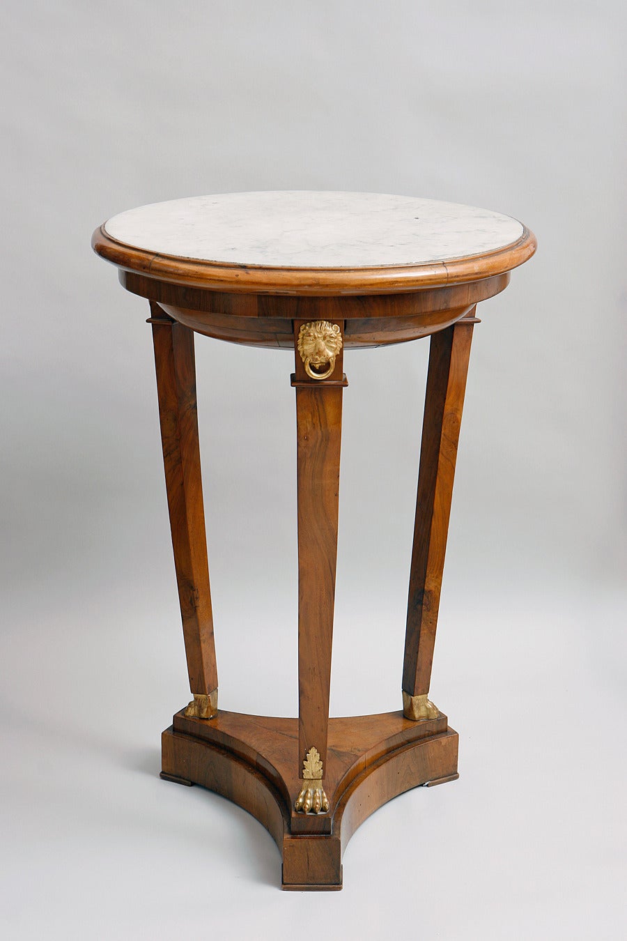 Nutwood, white marble top, gilt parts and applications. 
Three tapering legs with lion feets and heads uprising from a concave tripatited plinth. A very elegant and early empire furniture.

Compare with an Italian table from the Museo Napoleonico in