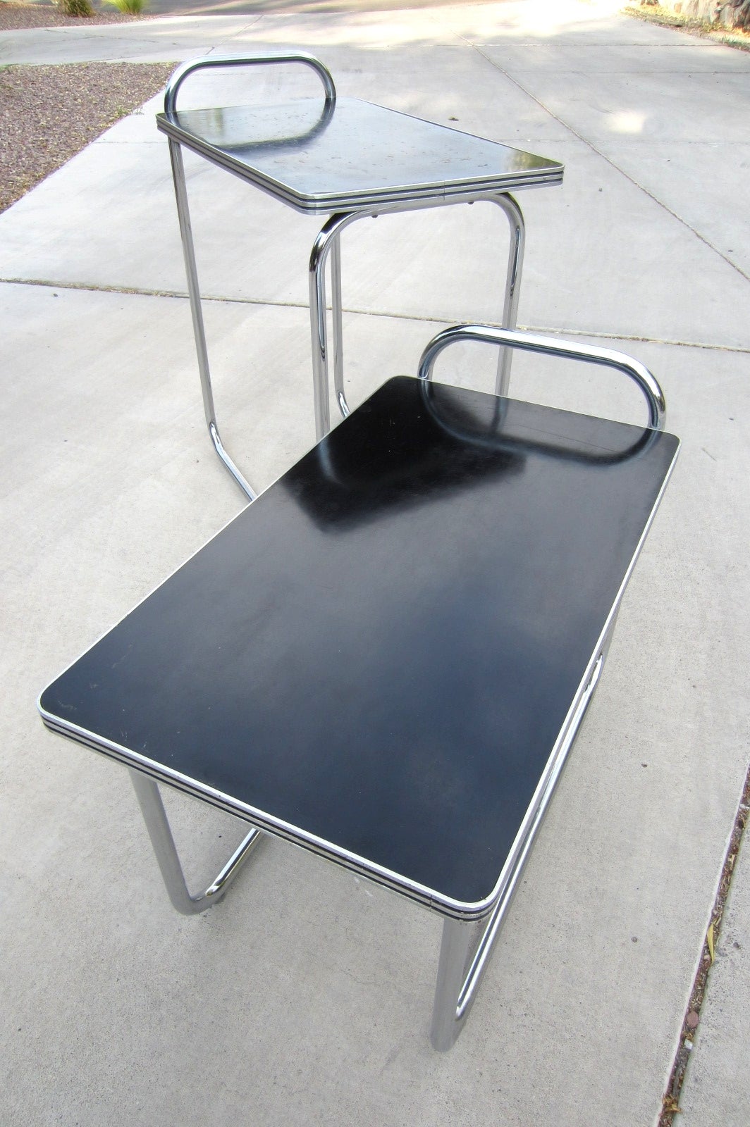 Beautiful set of two occasional service tables designed by Gilbert Rohde for Troy Sunshade Products.
Chrome plated seamless steel tubes with black lacquer tops surrounded in streamline design aluminum trim. Taller has original label as pictured.