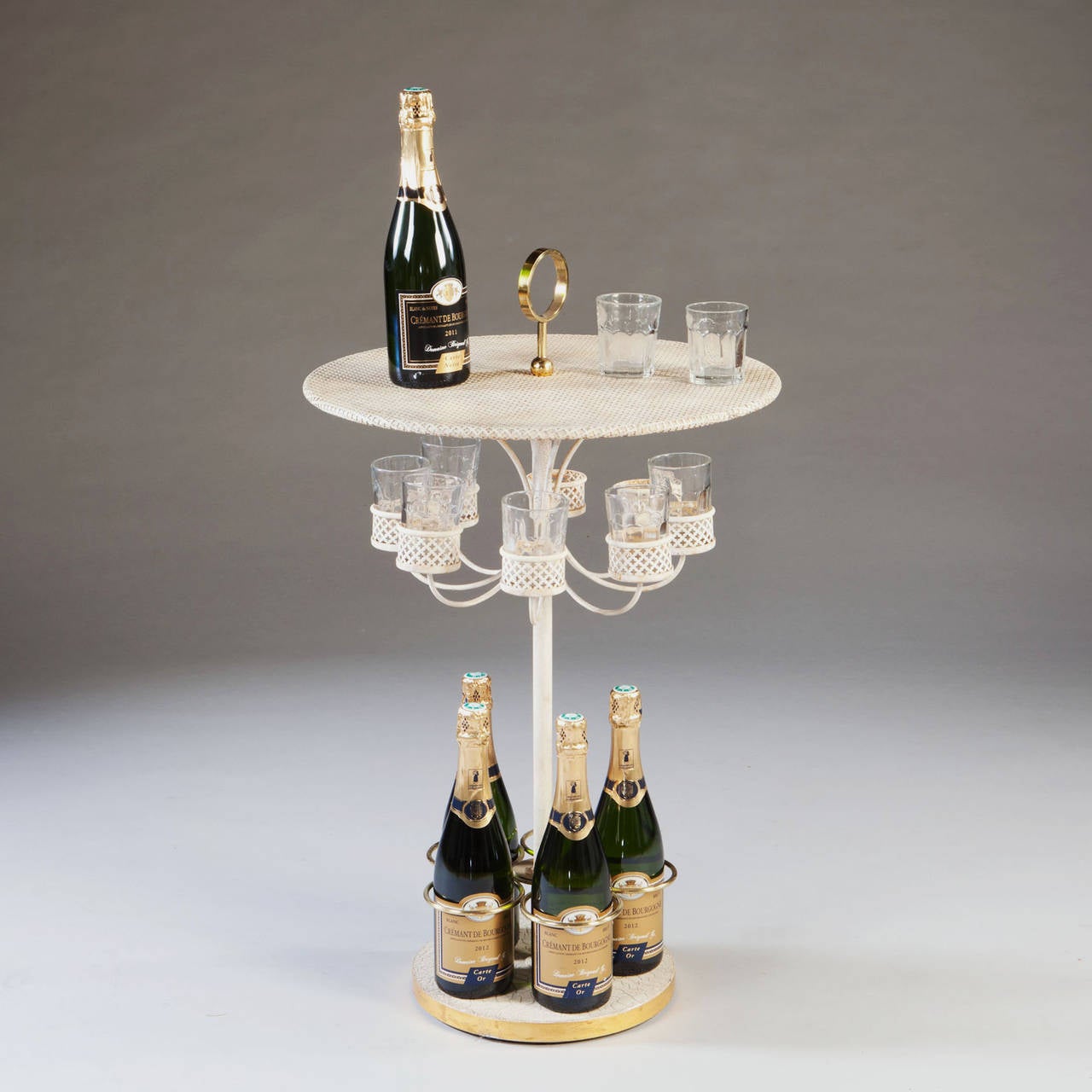 Pierced sheet metal is employed to create the circular top and the eight glass holders, the top having a brass carrying handle. At the base there are five brass rings to retain bottles and the whole stands on a weighted circular plinth