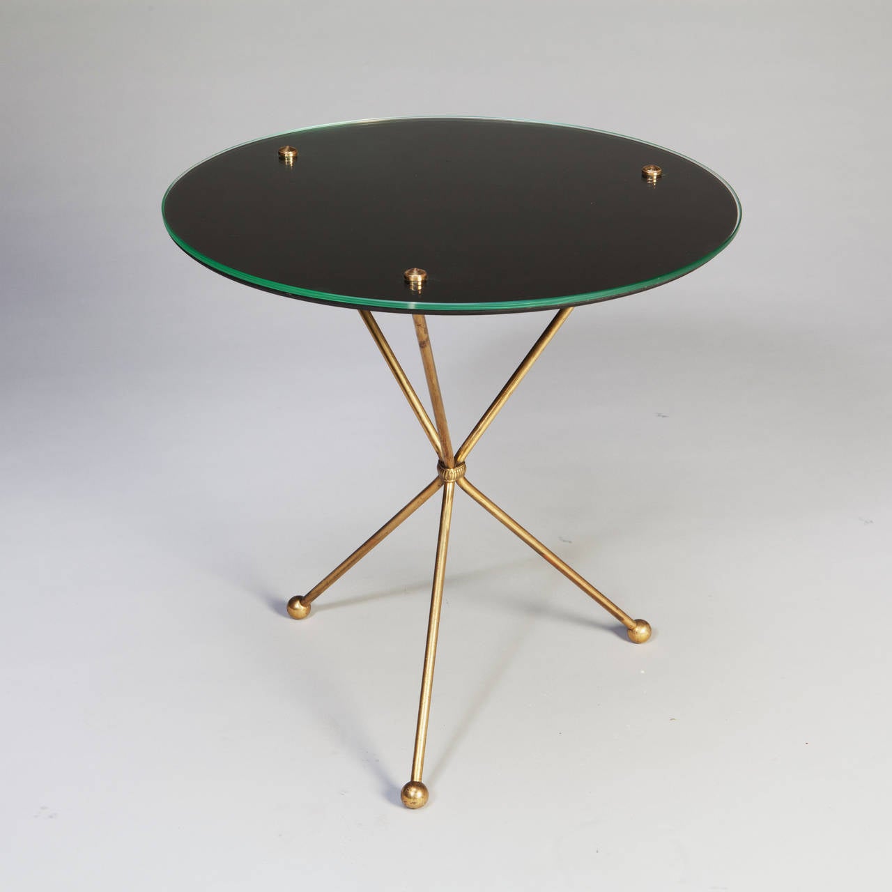 A pair of Mid-Century brass tripod end tables. The glass and black vinyl tops are held in situ by brass studs. The legs are cinched by a reeded strap. After the design of Jean Michel Frank.