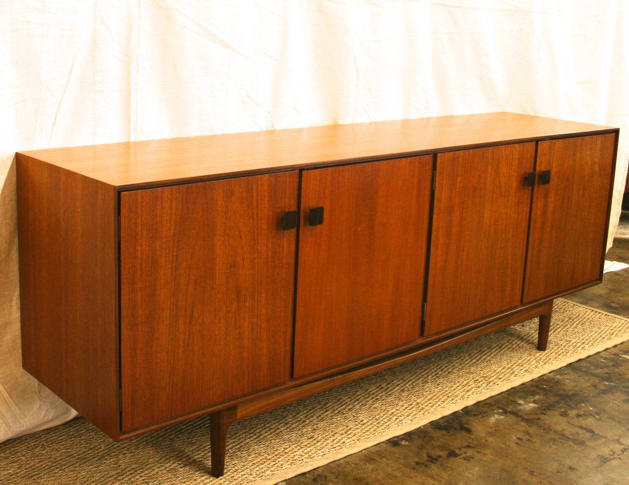 Long, stately teak credenza in the style of Ib Kofod-Larsen for G Plan. Prime example of minimal Mid-Century Scandinavian style. Four compartments, each with an adjustable shelf. Doors feature square pulls and minimal brass hardware, and close