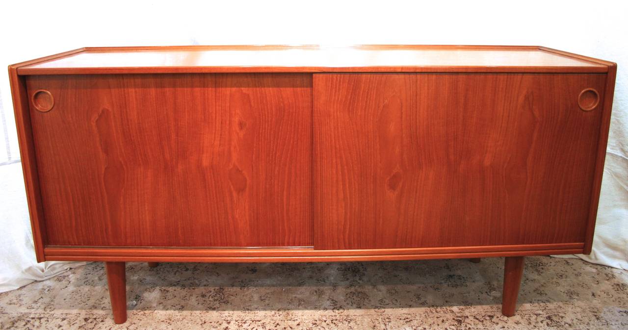 Compact teak credenza with sliding doors, circa 1960. Right side compartment features large storage space with one shelf which extends into left side. Left side also features two shallow drawers, lined in felt, for storage of small delicate items.