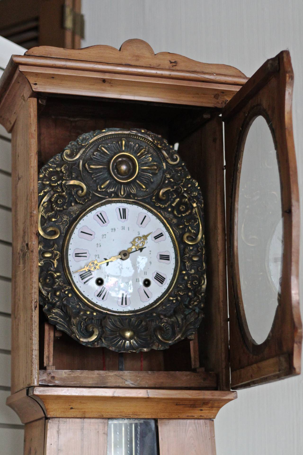 Long case clock with floral brass surround on enamel face.  Faint floral art along bottom section. Eight day repeating chimes at two minutes past the hour and at the half hour.