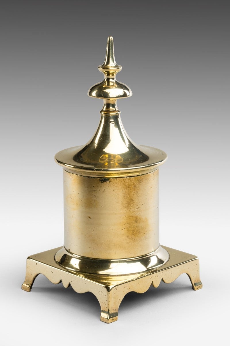 The removable cover topped by a spire finial, the canister-form body with concave base moulding, raised on a square bracket plinth with ogee-shaped friezes. A quotation from 'Domestic Metalwork' sums up the use of brass for making objects in daily