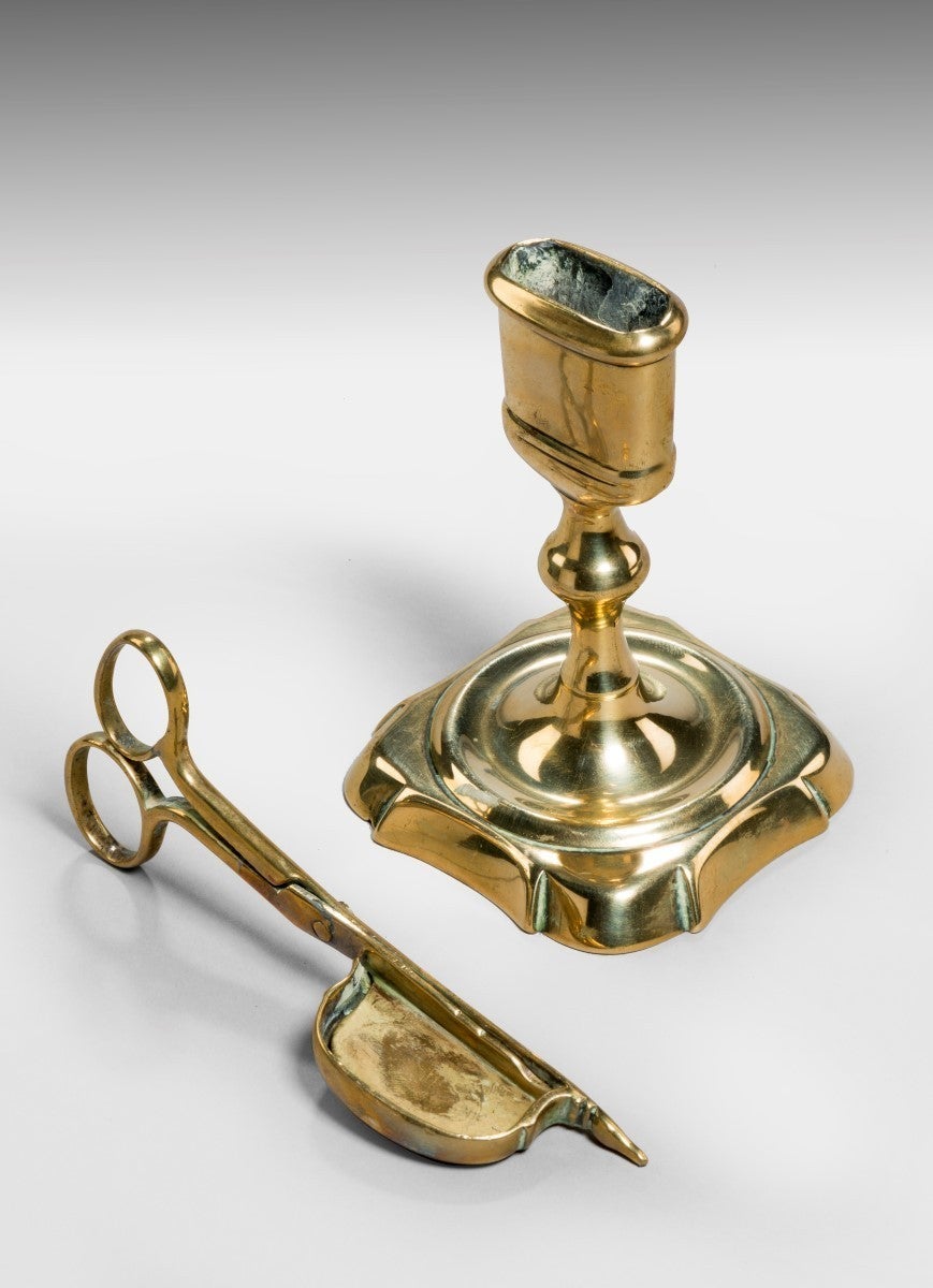 A brass candle snuffer with scissor-like handles and a pointed tip, which is accommodated vertically into the stand. With a square based stand. There are two holes in the bottom of the stand so that the point on the snuffers can go in either side.