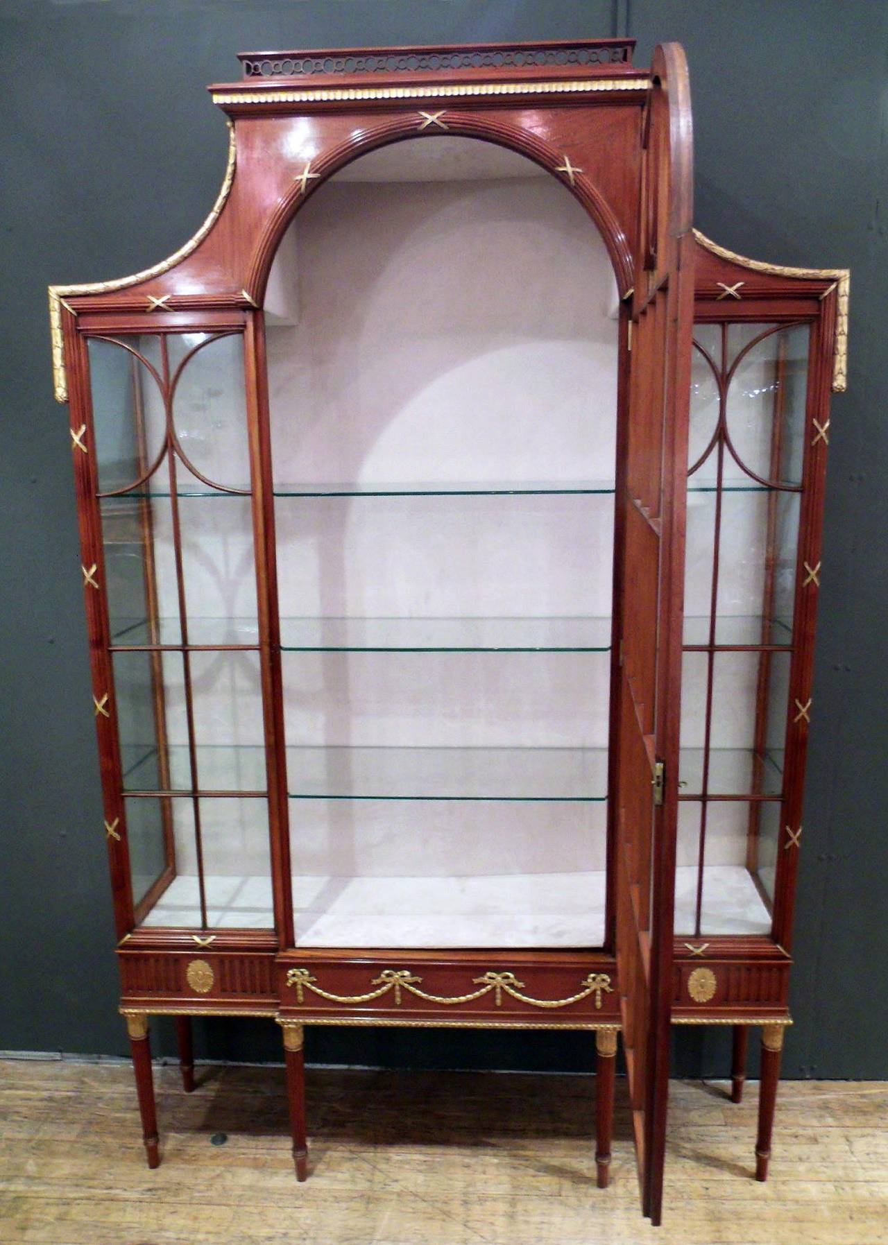 This Edwardian satinwood and parcel-gilt display or China cabinet was made by Maple and Co. of London and stands on six tapered, reeded legs below a breakfront frieze with gilt swag and ribbon decoration. The vitrine features an astragal glazed