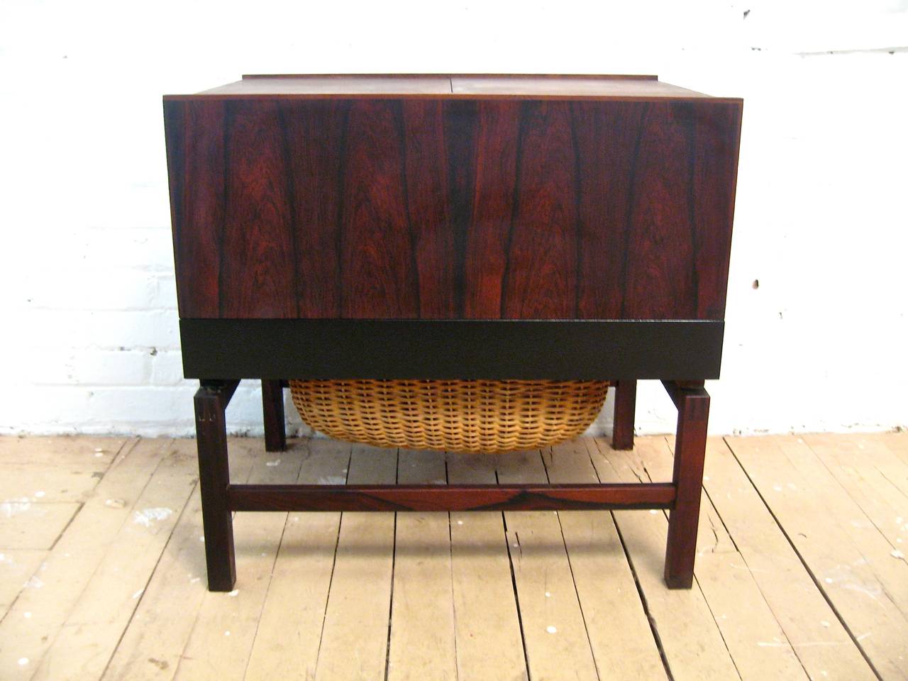 Wonderfully figured rosewood grain enhances this work table/sewing stand.  This piece features two slide down doors that hide the interior storage compartment and a black lacquered bottom drawer with wicker basket (meant for storing yarn, etc.). 