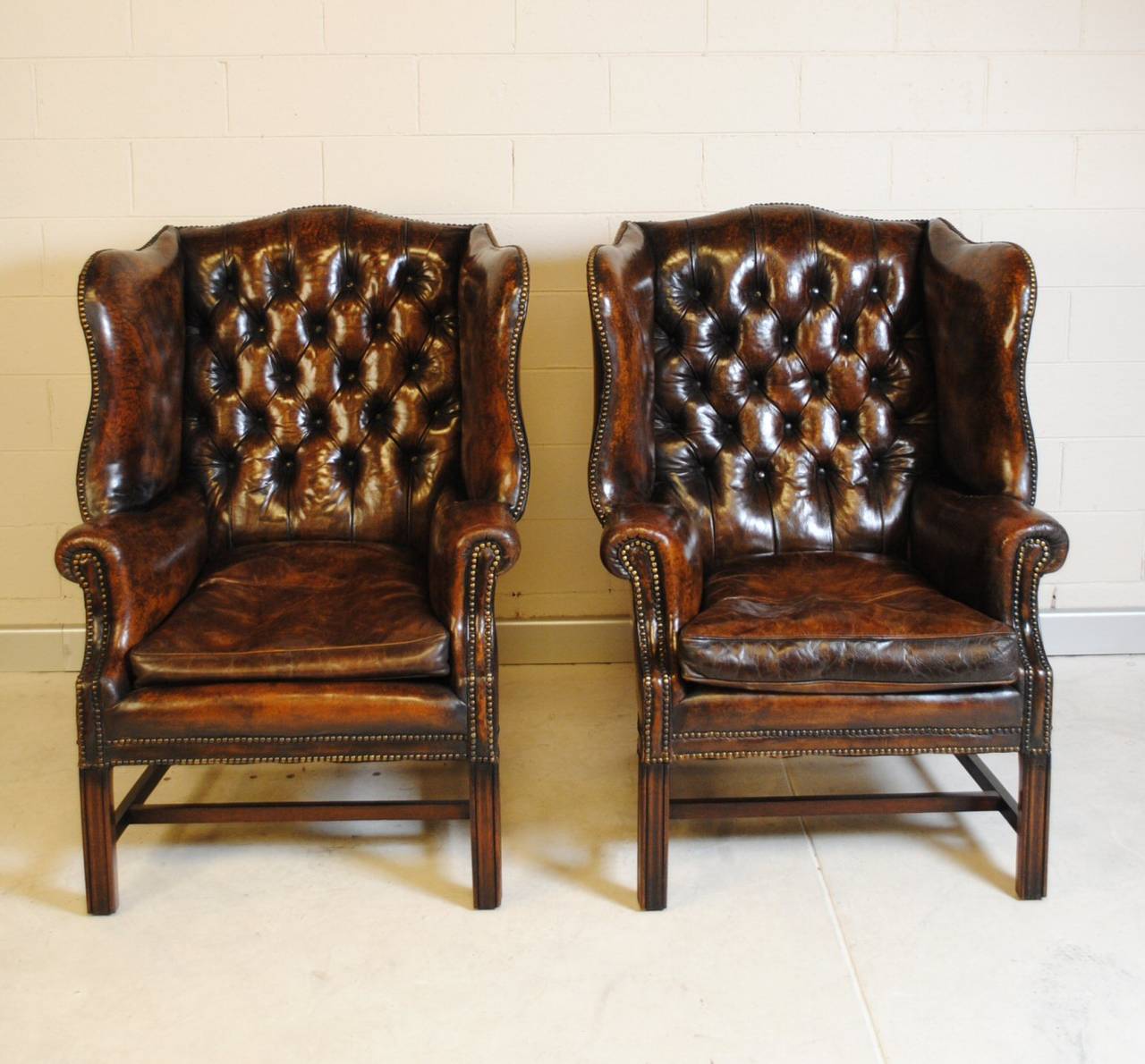 Pair of English wingback leather chairs with button tufted backs, brass studs and sturdy 'H' frame stretcher bases. A classic pair of chairs with a deep, rich patination perfect for the study. Dating from the early 1900s.