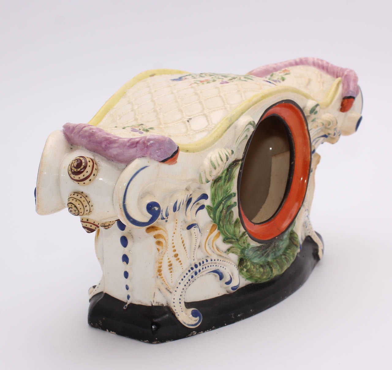 Remarkable English pearlware pocket watch stand, modelled as a wide mantel clock, a large scroll to either side decorated with snail shell knops, the upper surface with a net and painted flowers, the opening for the watch framed by a green garland