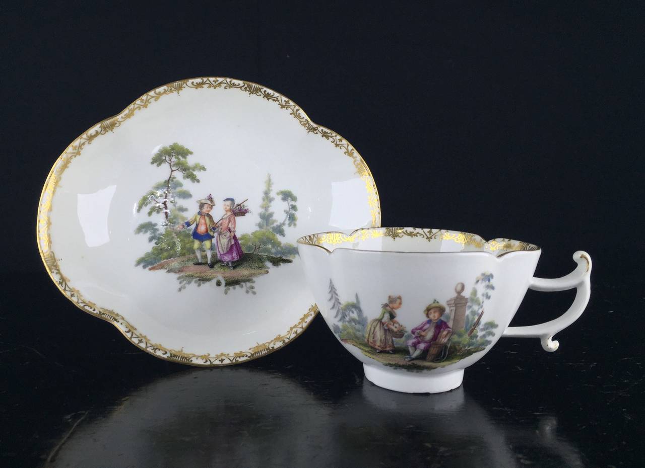 Rare Meissen quatrefoil shape cup and saucer with elegant handle, finely painted with children in landscapes, one side of the cup with a boy showing a girl his empty grape basket, while on the other side he plays a pipe to her, his basket being full