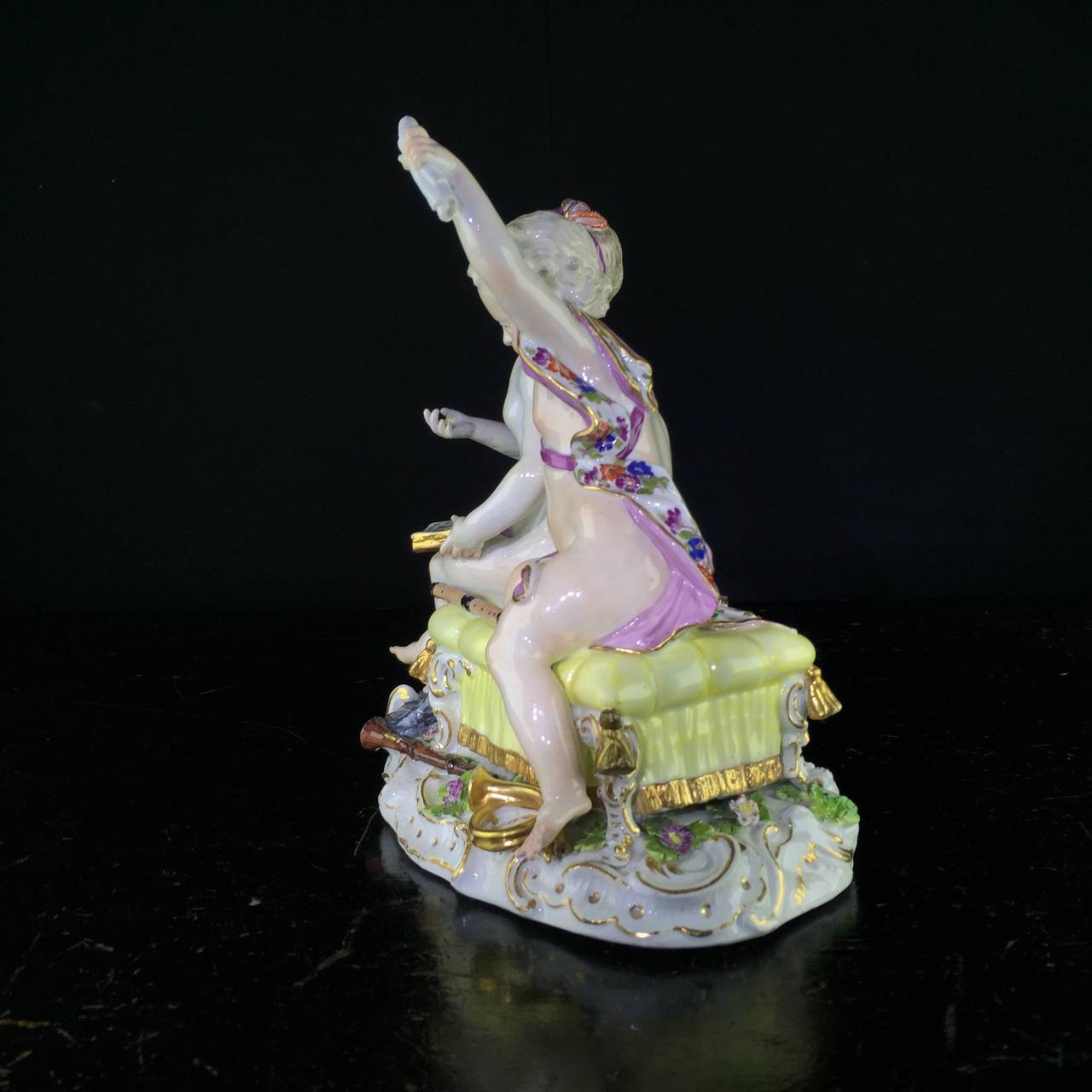 Meissen figure group of musical children, with a semi-clad girl & boy seated in a large yellow upholstered stool with gilt fringe and tassels, singing from an open book with musical notes and the title ‘Aria.’, with various instruments scattered