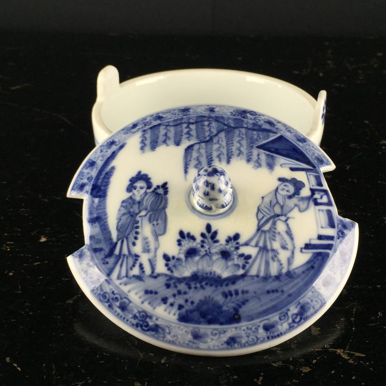 Meissen butter tub, painted in underglaze blue with chinoiserie scenes, the cover with two oriental ladies in a garden, the base with a continuous landscape, the knop modelled as an artichoke and picked out in blue. 
Impressed five, 
early crossed