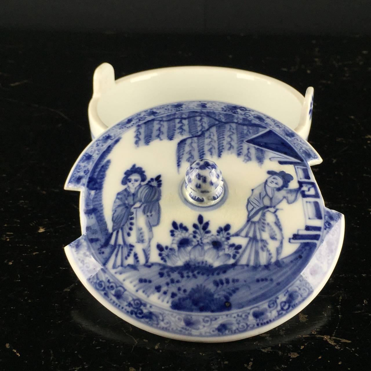 Meissener Butterfass, in Unterglasurblau bemalt mit Chinoiserie-Szenen, der Deckel mit zwei orientalischen Damen in einem Garten, der Boden mit einer durchgehenden Landschaft, der Knauf als Artischocke modelliert und blau gepickt. 
Beeindruckt