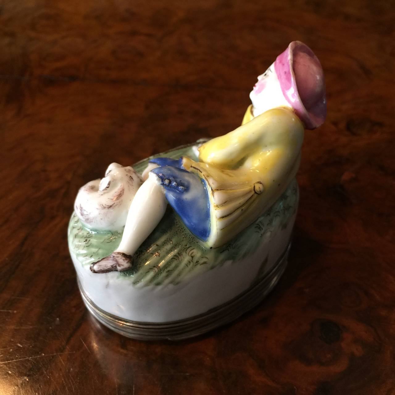 Mennecy snuff box of oval shape, modeled with a reclining shepherd and his dog to the lid, wearing a bright yellow coat, blue trousers, and a pink hat, the lid moulded & painted with flowers inside & out.   

  Silver mounts hallmarked Paris 1762 