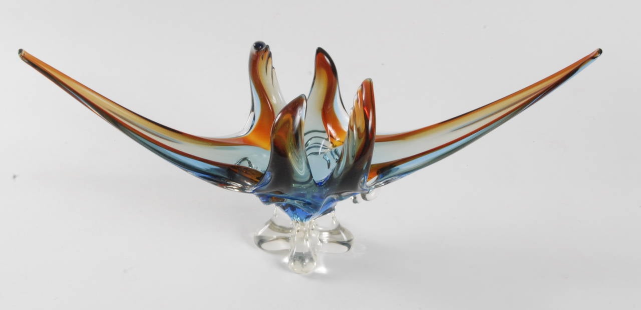 A very large Murano glass table center with blue and orange color through a clear body, clear four footed base.
Long sweeping gently curving tendrils with four shorter and heavily curved central arms creating a closed central section to give height