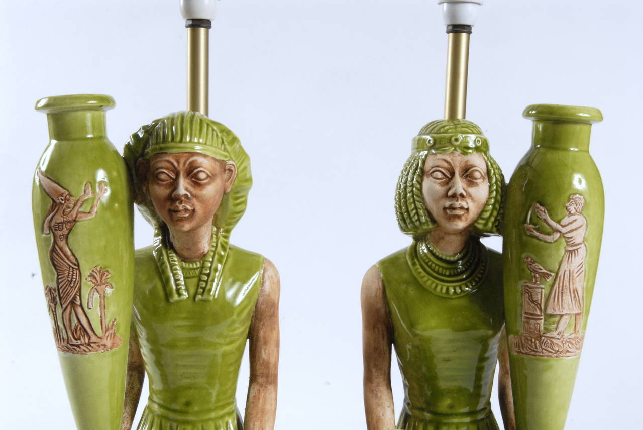 A highly original pair of Italian ceramic lamp bases depicting an Ancient Egyptian couple, a Pharaoh and his consort, both kneeling on brown detailed bases and holding large oil vases. Made as a mirror pair and having naturalistic skin tones.