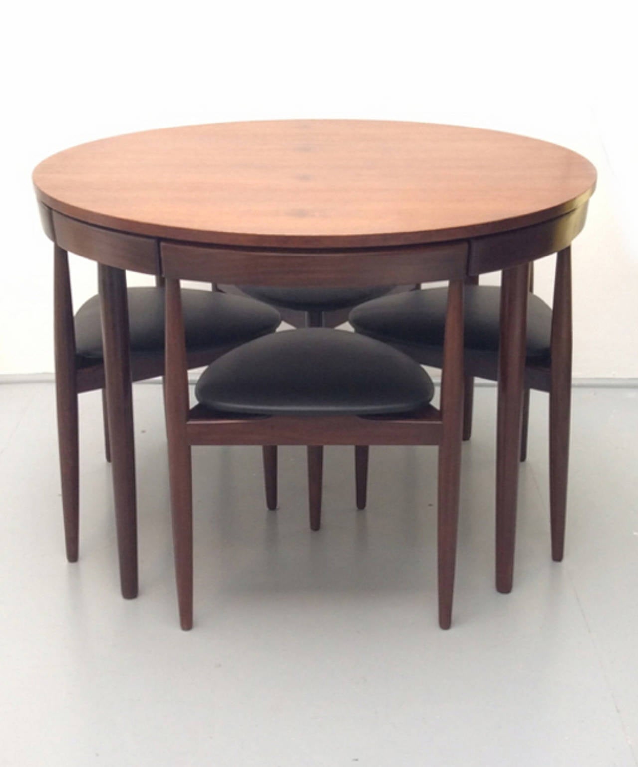 The teak and afromosia suite comprises four three-legged chairs which fit neatly into position in the apron of the table top. The chairs have been newly re-upholstered in soft black leather, and pieces are labelled.  Table legs unscrew for