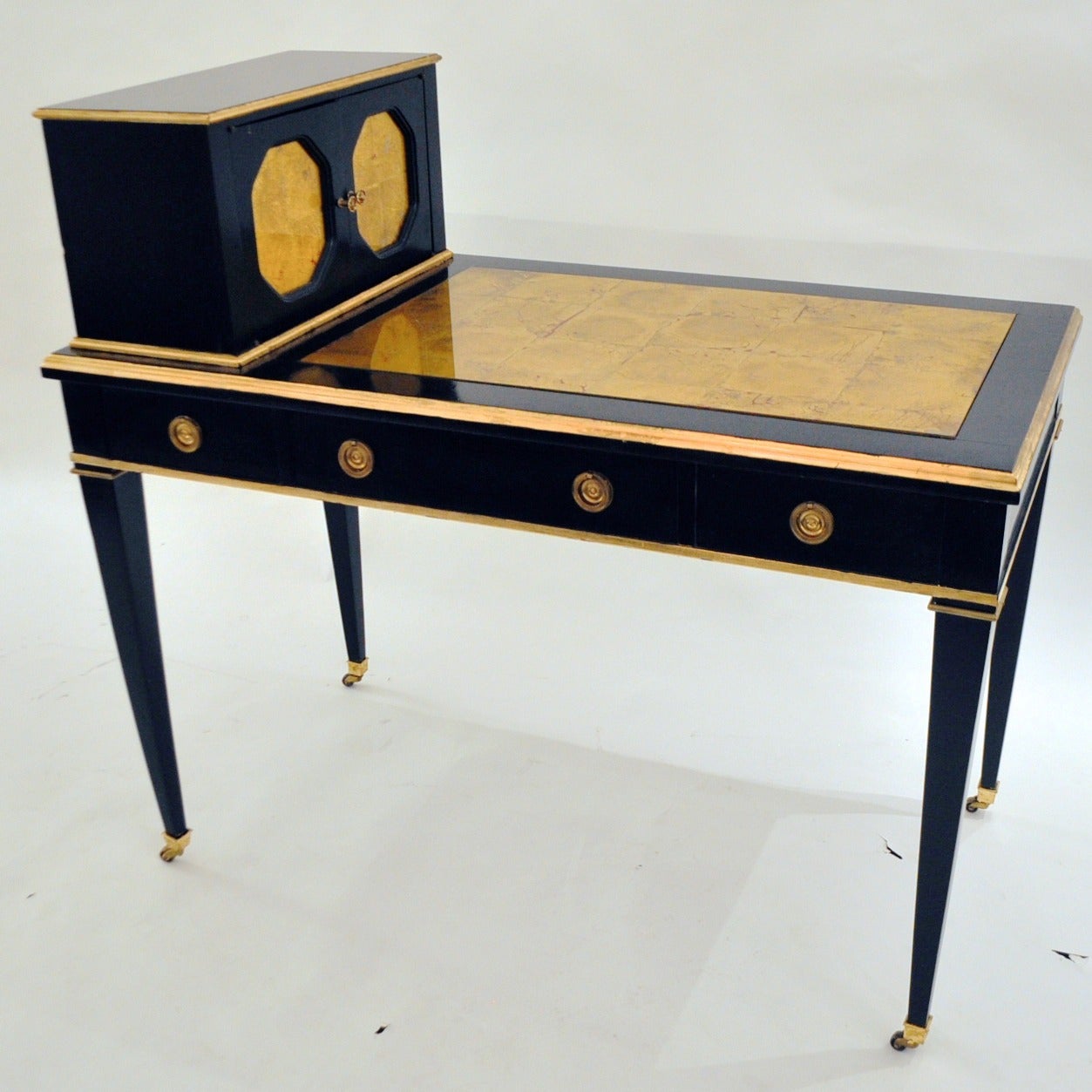 Desk is done in ebony with gold leaf mirrored door fronts and desk top writing area. It has brass drawer pulls knobs and castors.