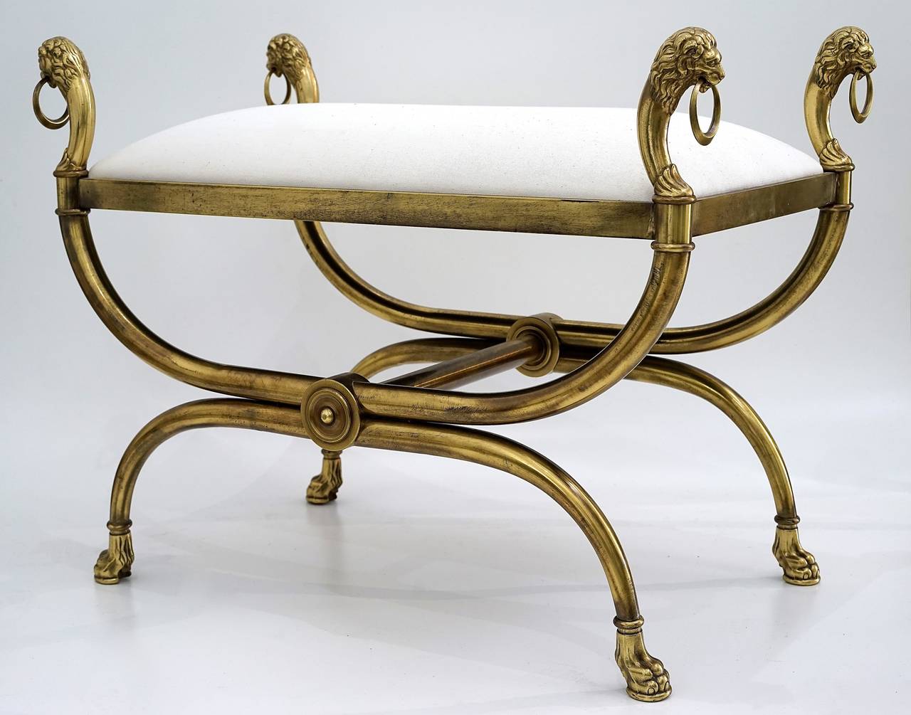 This Mid-Century Curule-Style bench has an elongated X-Frame with lion-paw feet, lion-heads with undulating-manes and each grasping a crescent moon- shaped ring. 

The frame is an antique brass/bronze coloration.

The slip-seat is newly