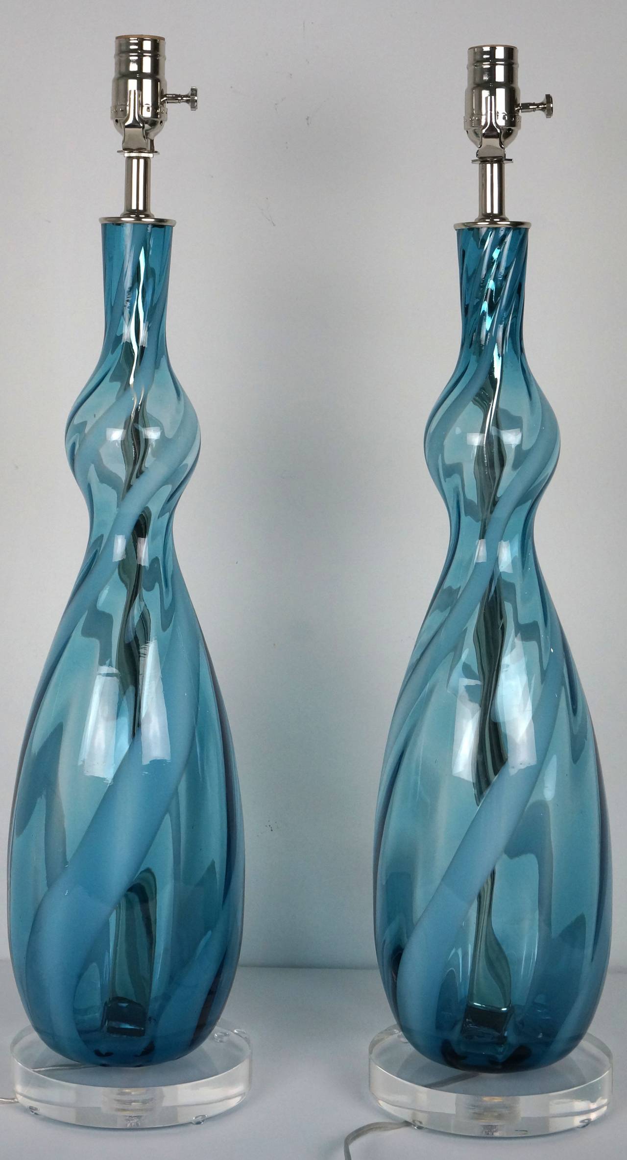 This beautiful pair of Murano glass lamps were created in the 1960s.

The vase form with a bulbous neck detail and swirling ribbons in colors of clear, peacock-blue and white make for a very light and airy composition. 

These lamps have been